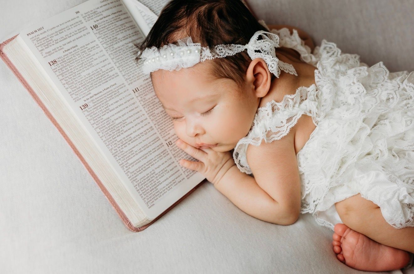 Peaceful baby girl in white lace outfit sleeping beside an open Bible with floral headband.
