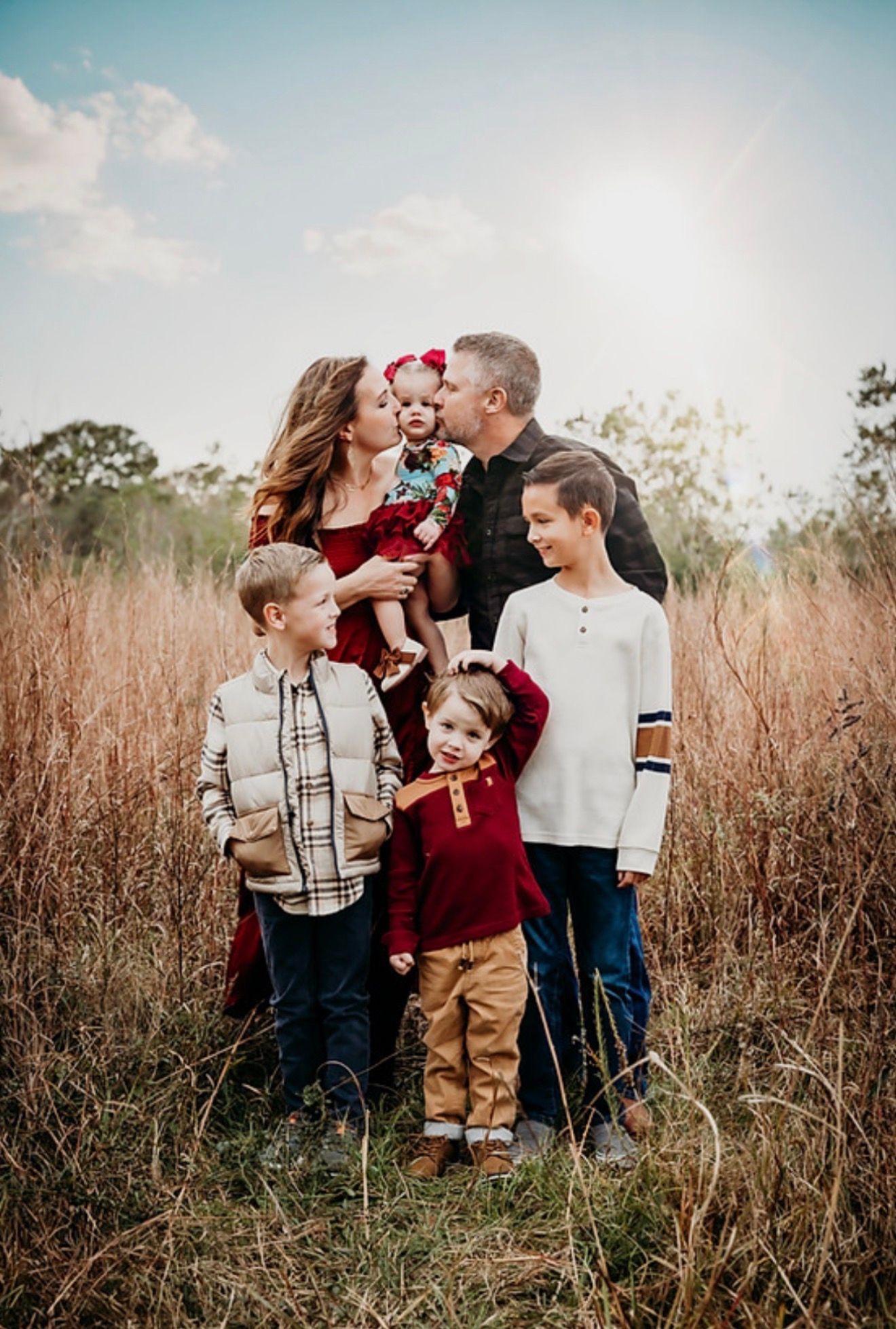 Family of five posing in golden field, parents kissing baby girl while three children smile, wearing coordinated fall outfits.