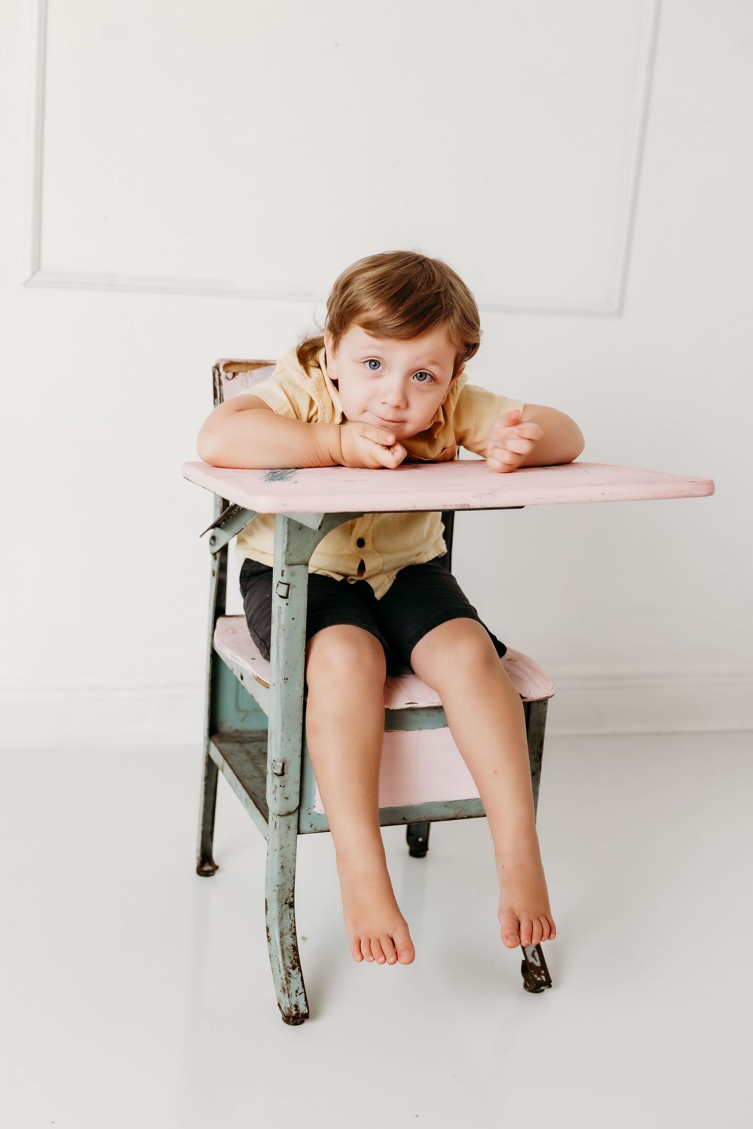A young child sits barefoot at a vintage pink school desk, resting their chin on their arms with a contemplative expression.