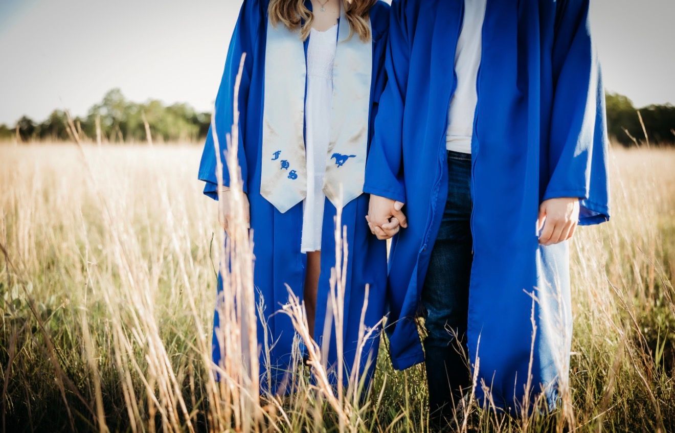 Two graduates in blue gowns hold hands while standing in tall golden grass field.
