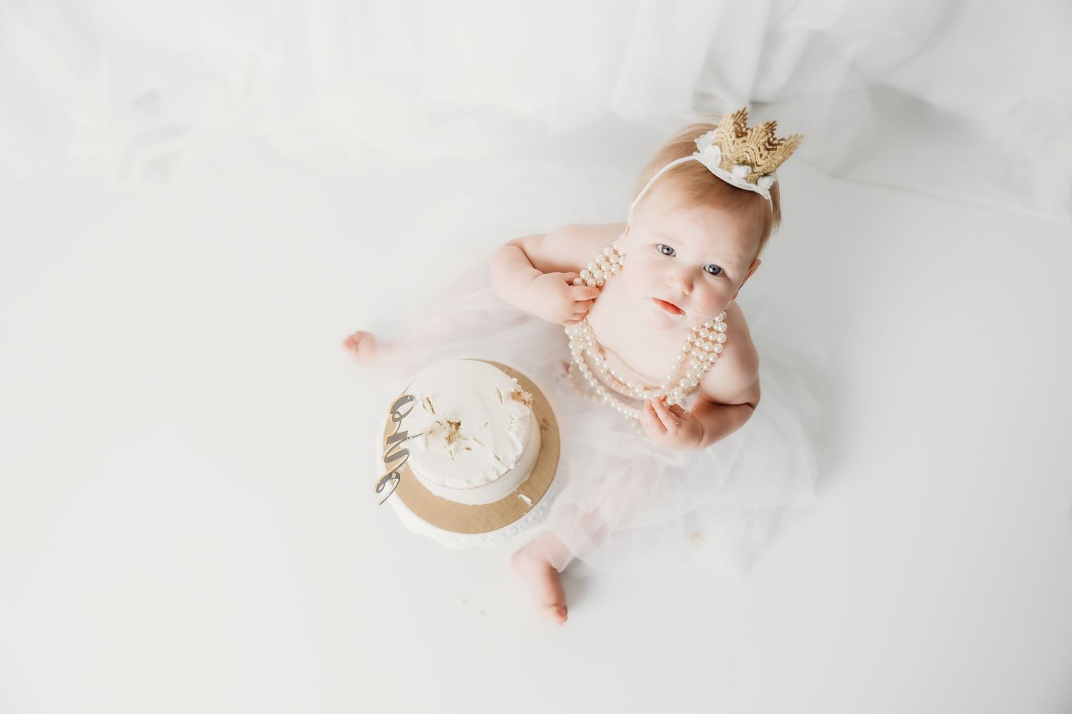 Baby girl in white tutu and gold crown sits on white floor with first birthday smash cake.