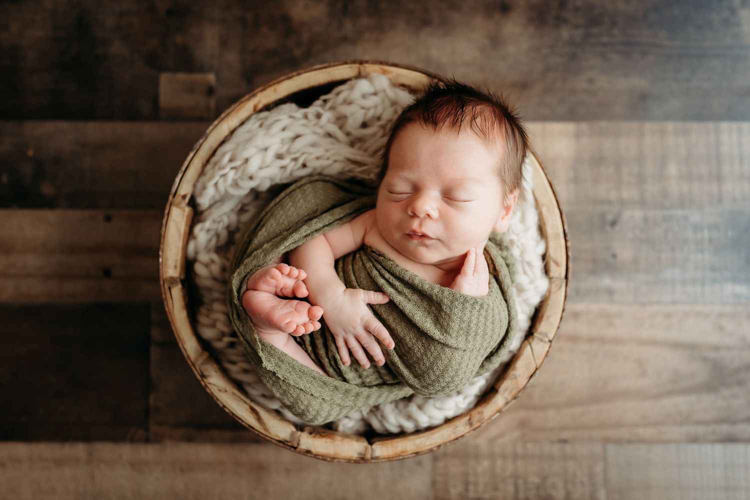 Sleeping newborn baby wrapped in green swaddle nestled in rustic wooden basket with cream knit blanket.