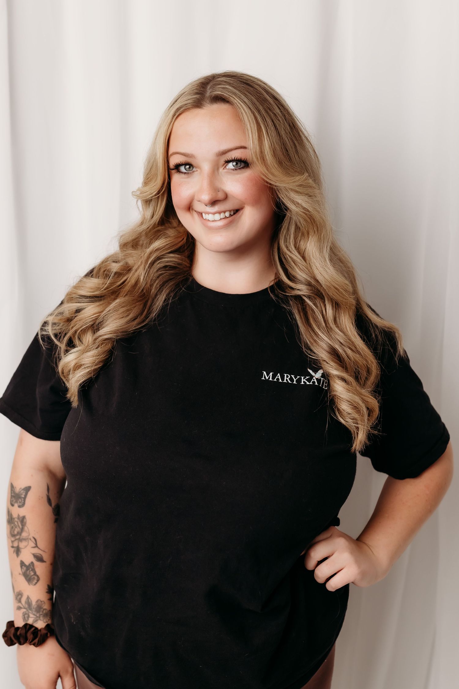 Smiling blonde woman in a black Mary Kay t-shirt with tattoos, posing confidently against a white backdrop.