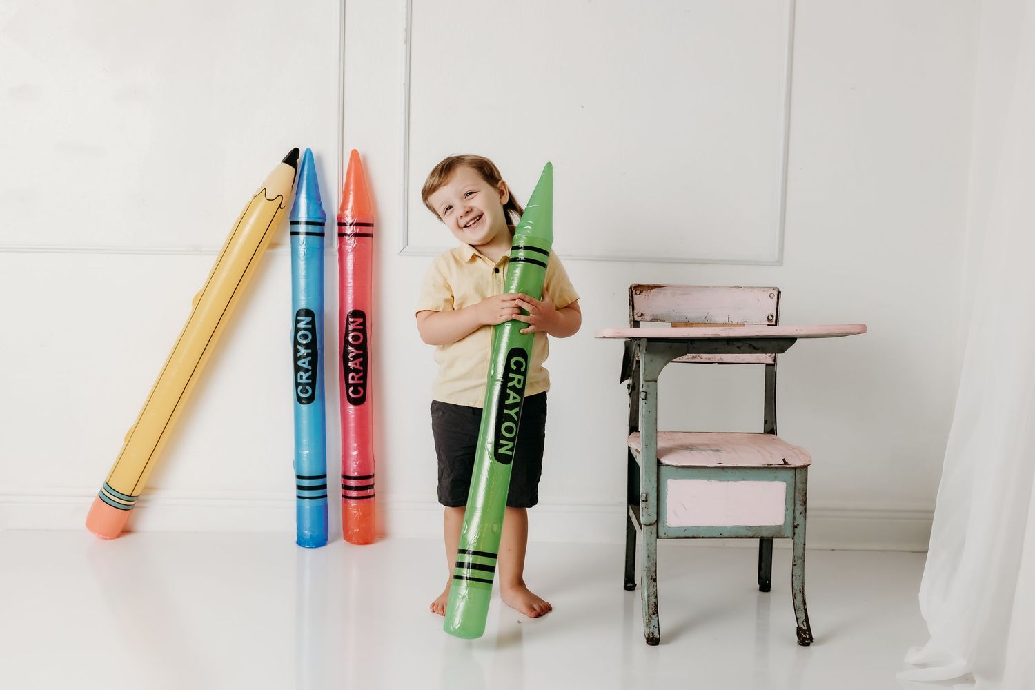 Smiling young boy holds oversized green crayon beside vintage pink desk and giant crayon props.