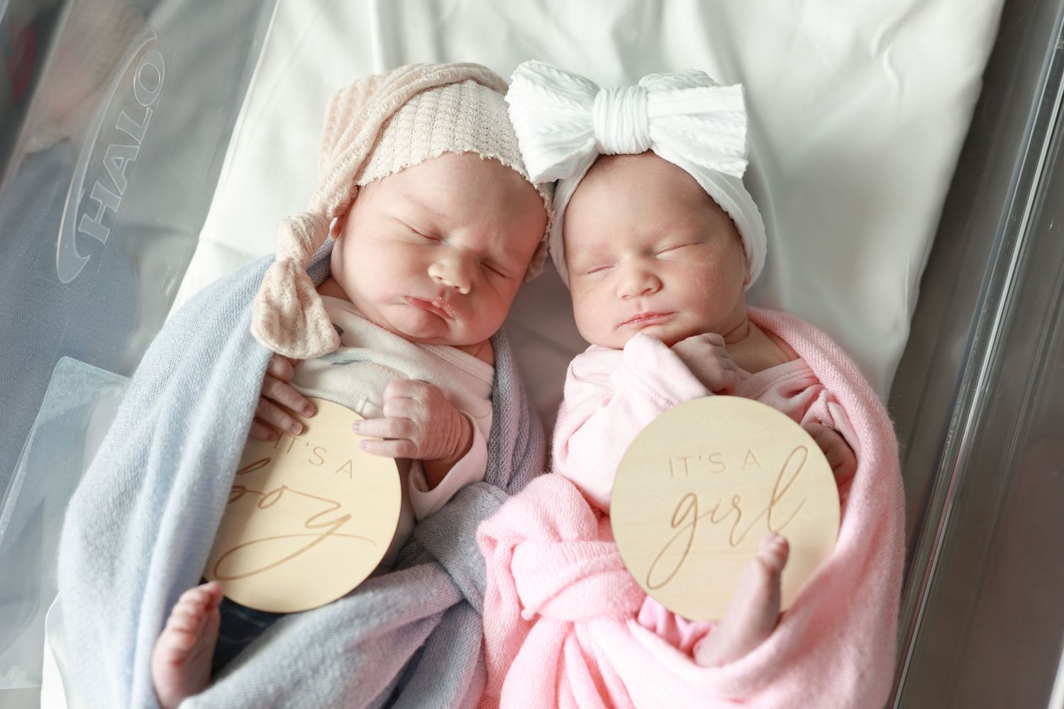 Twin newborn baby girls swaddled in pink and white blankets holding wooden girl announcement discs.