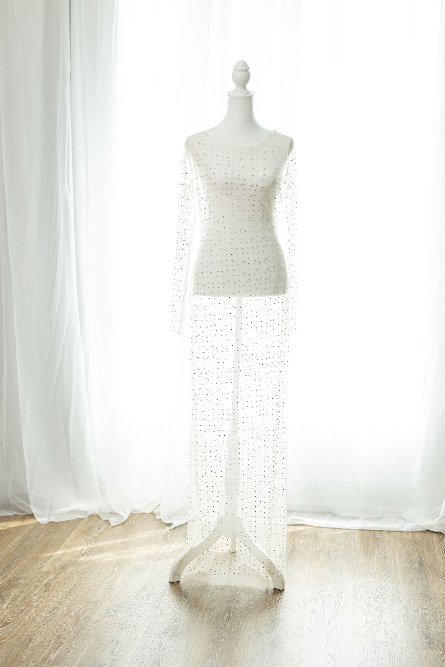 White beaded long sleeve gown displayed on a mannequin, floor-length with sheer embellished fabric, set against white curtains.
