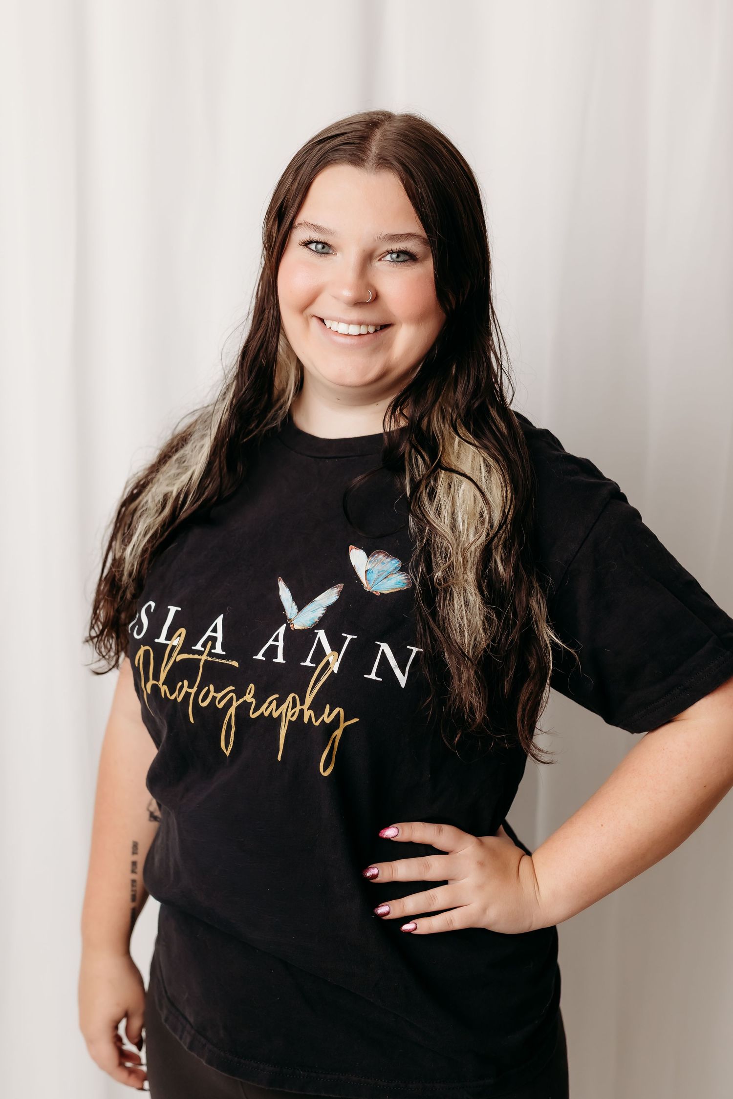 Smiling young woman wearing a black SLA Ann Photography t-shirt with butterfly design, posing against a white backdrop.