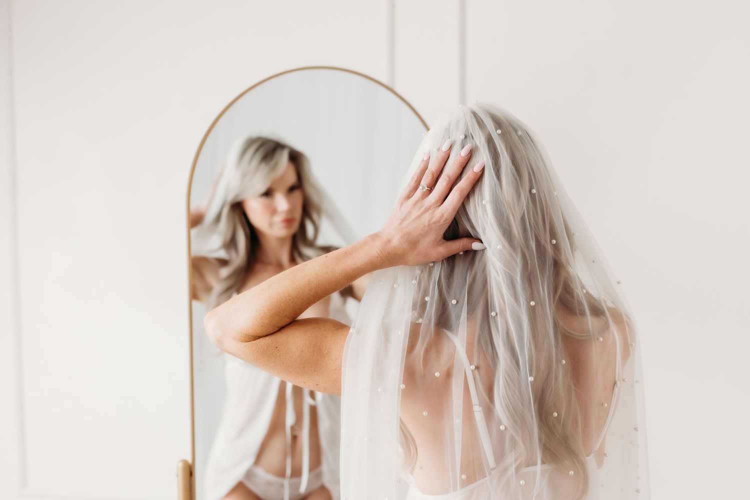 Bride wearing a pearl-adorned veil adjusts her hair while gazing at her reflection in an arched gold mirror.