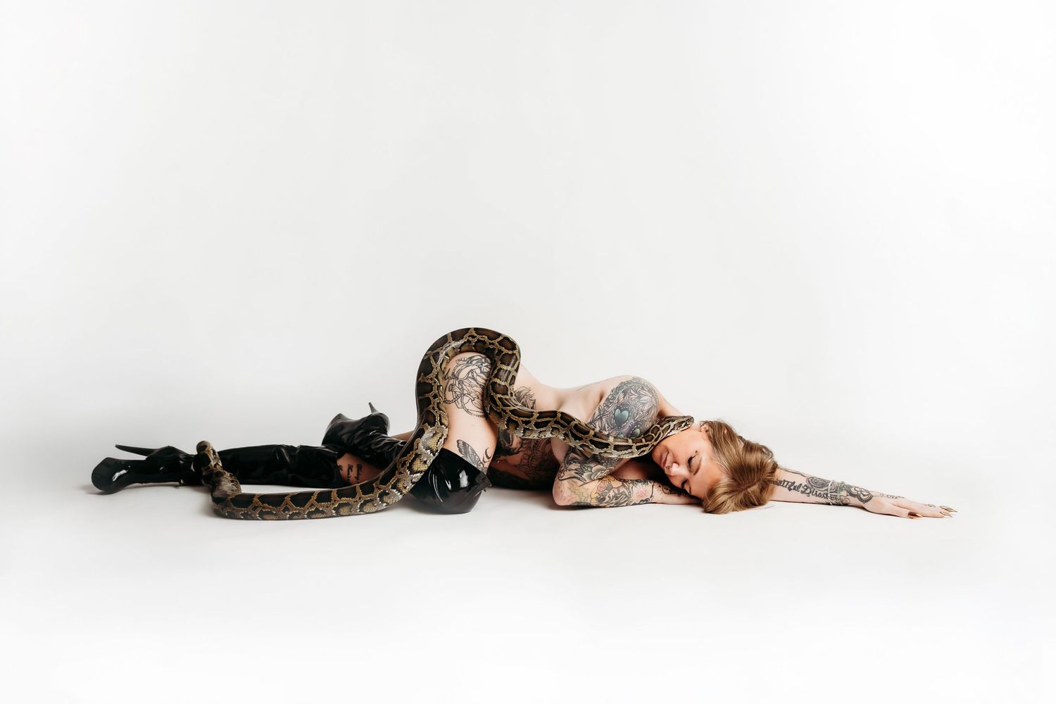 Tattooed woman in leopard print leggings poses with a large python snake on white background.