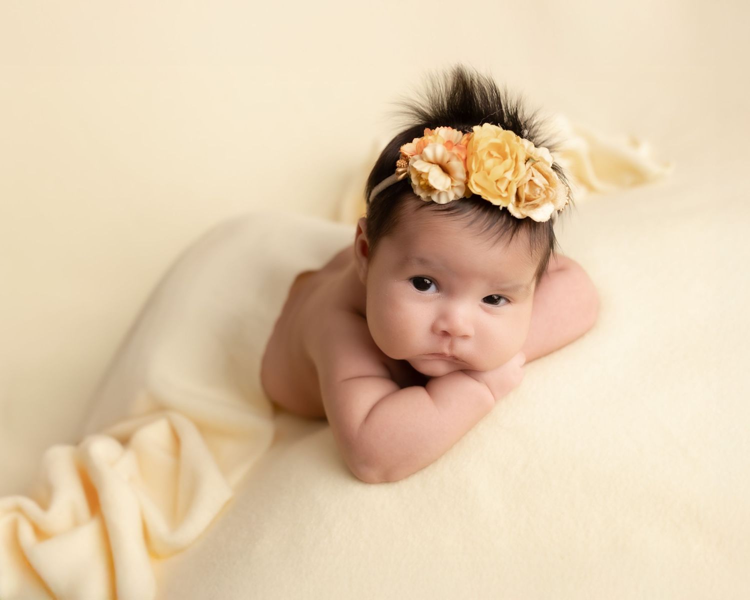 Newborn baby girl wearing a floral headband lying on a cream blanket in a professional newborn photography pose.