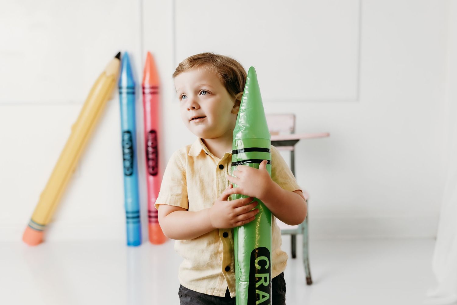 Curious toddler boy clutches giant green crayon prop in bright white photography studio setting.