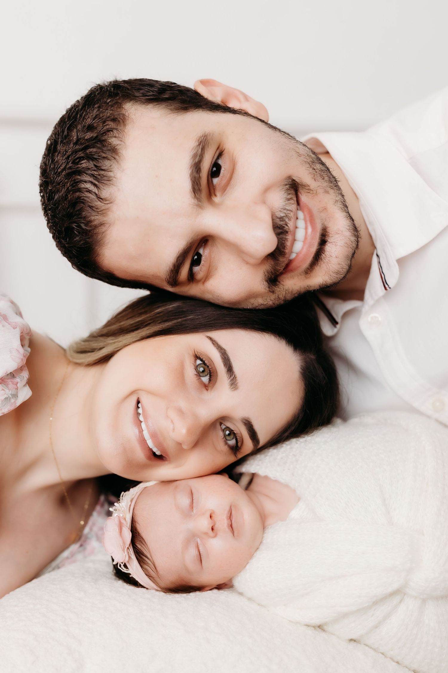 Smiling parents pose with sleeping newborn baby girl in white headband.