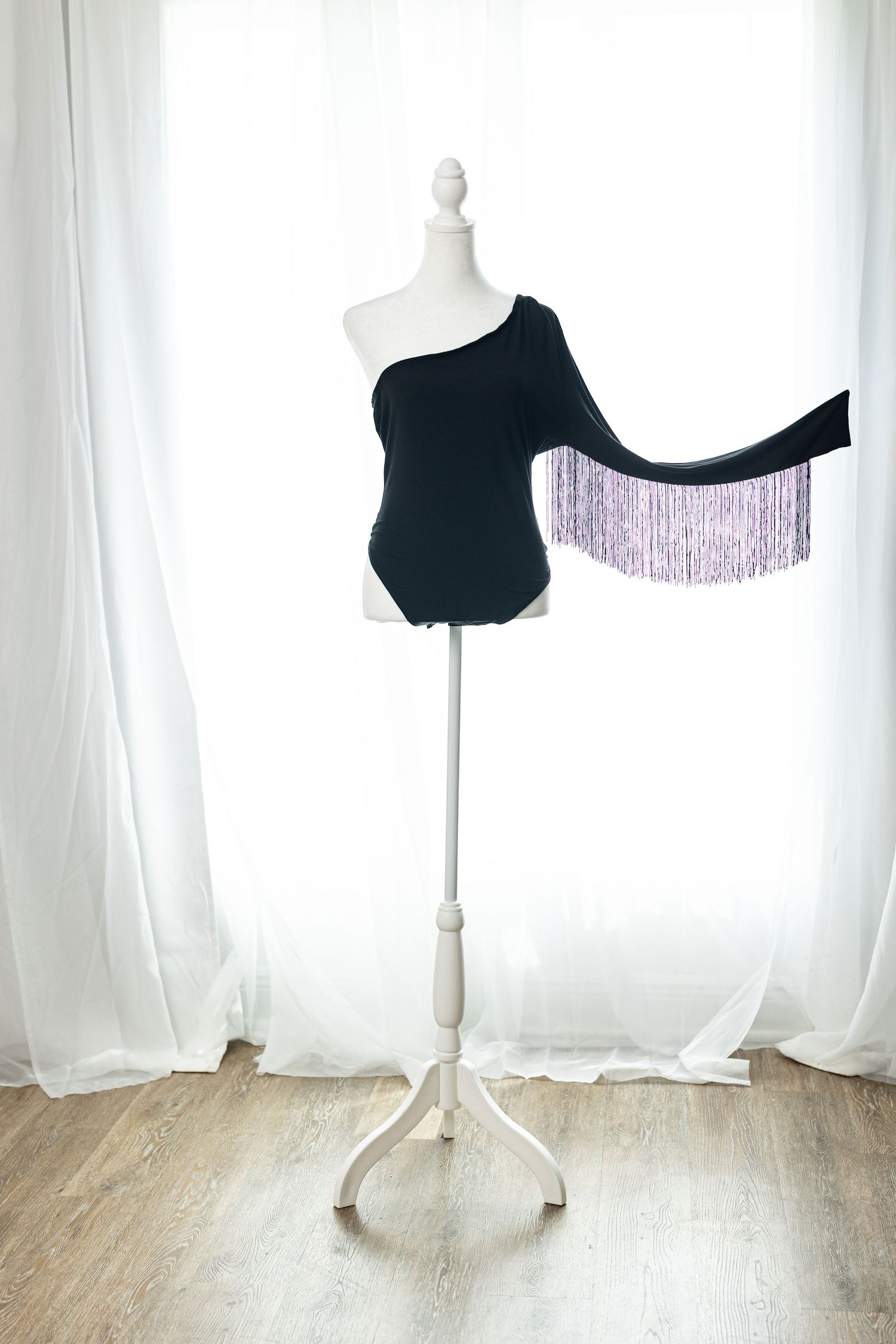 Black one-shoulder bodysuit with purple fringe detail displayed on a white mannequin stand against sheer curtains.