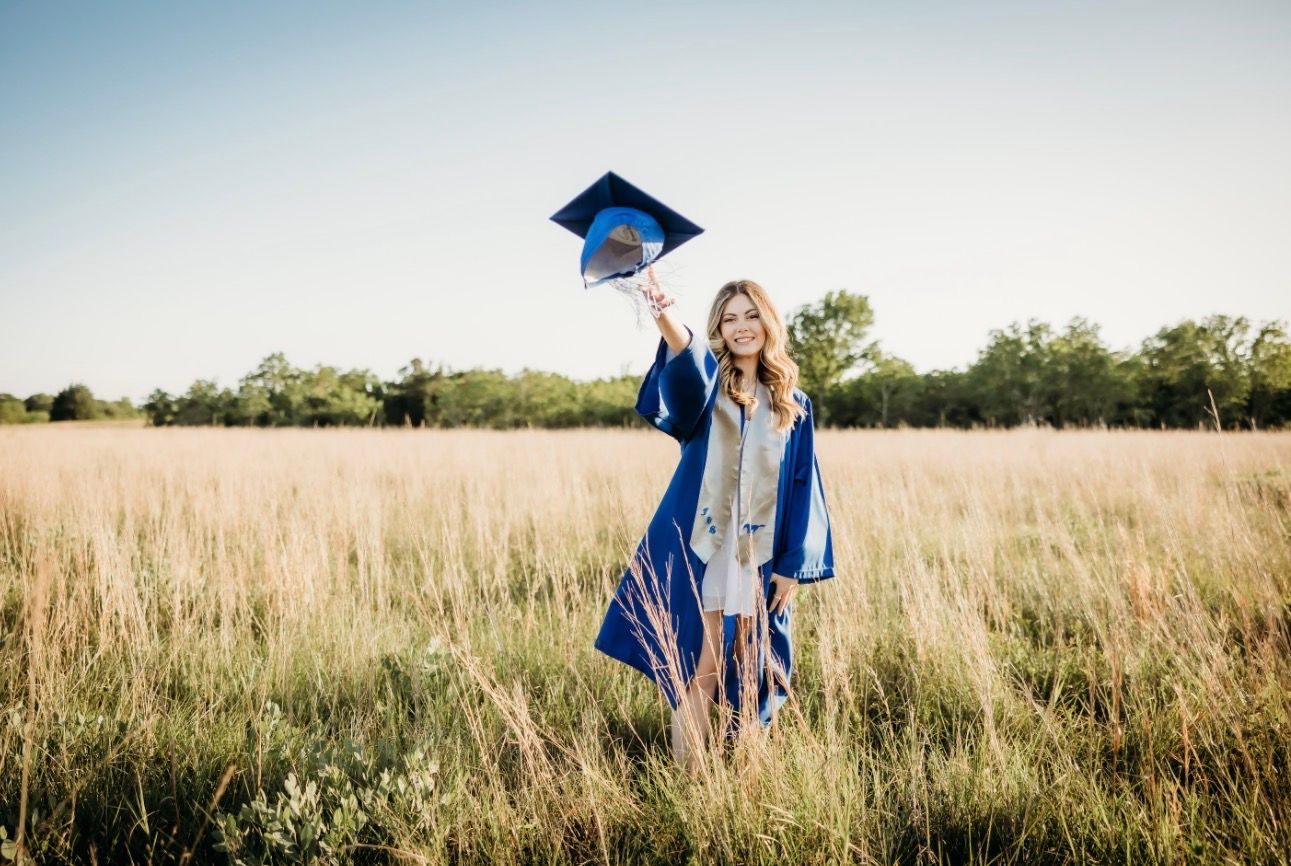 Graduate in blue cap and gown holding mortarboard up in sunny golden grass field.