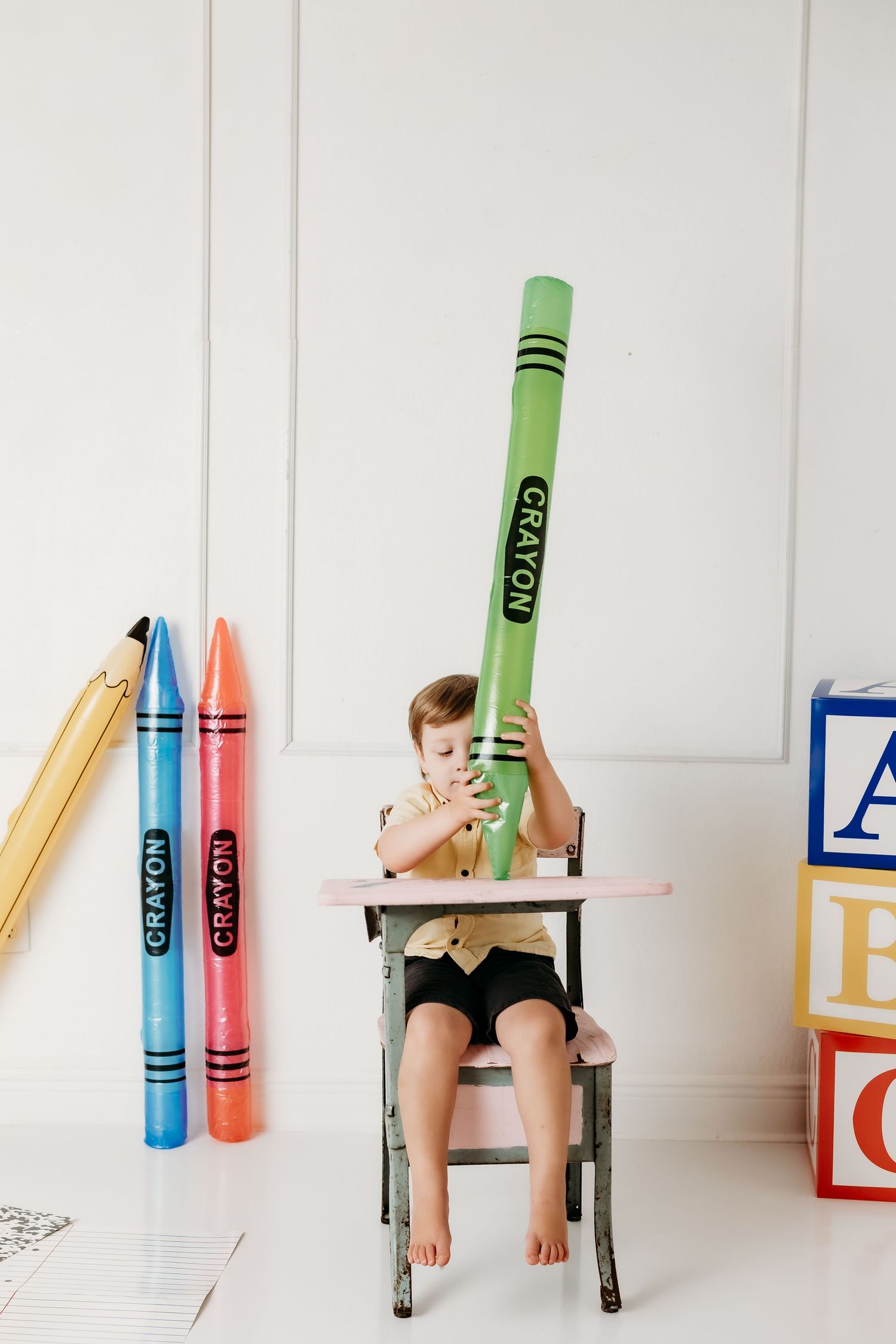 Young boy holds giant green crayon upright while seated at vintage pink desk with alphabet blocks.