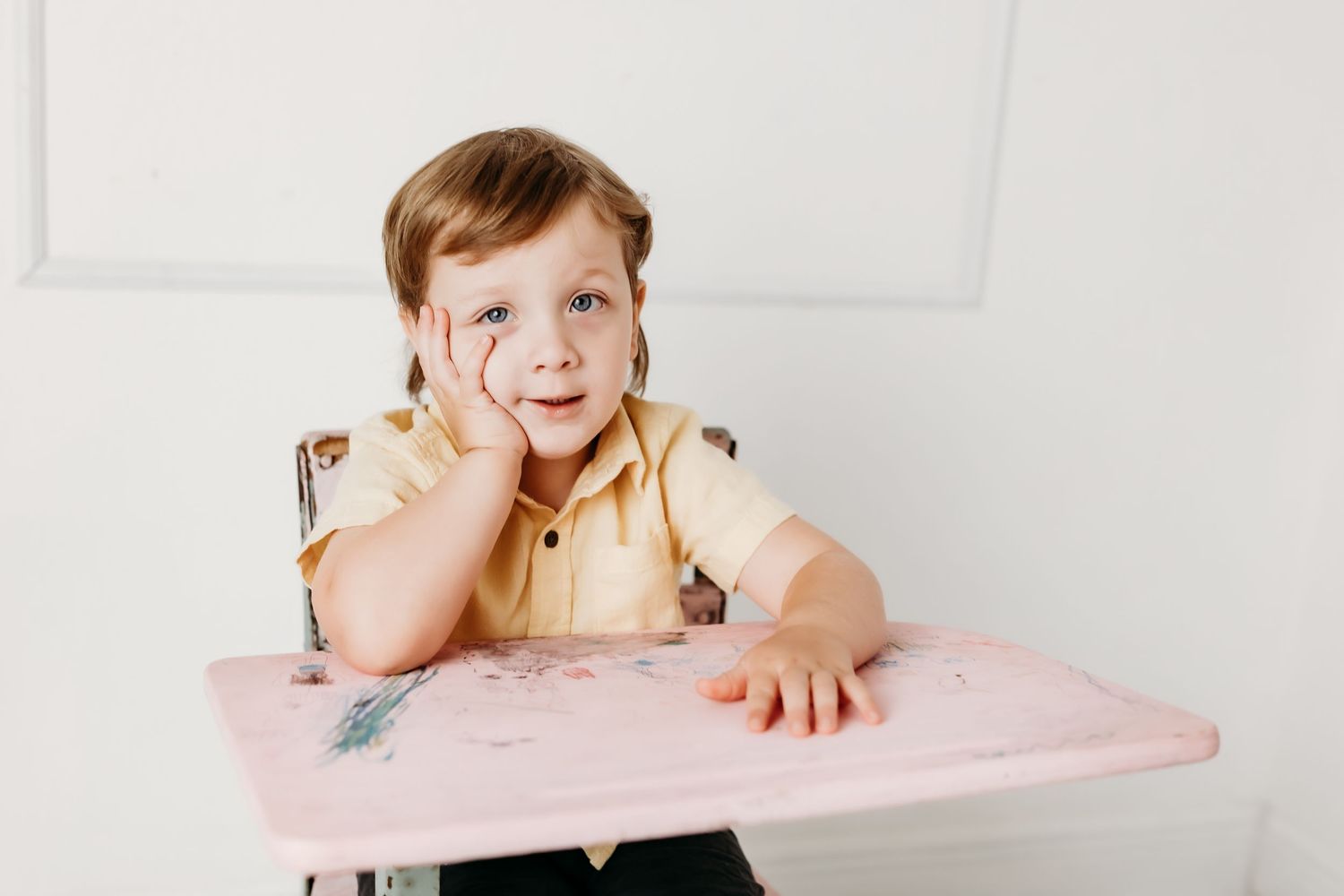 A young child in a yellow shirt sits at a vintage pink desk, resting one hand on their cheek with a cheerful expression.