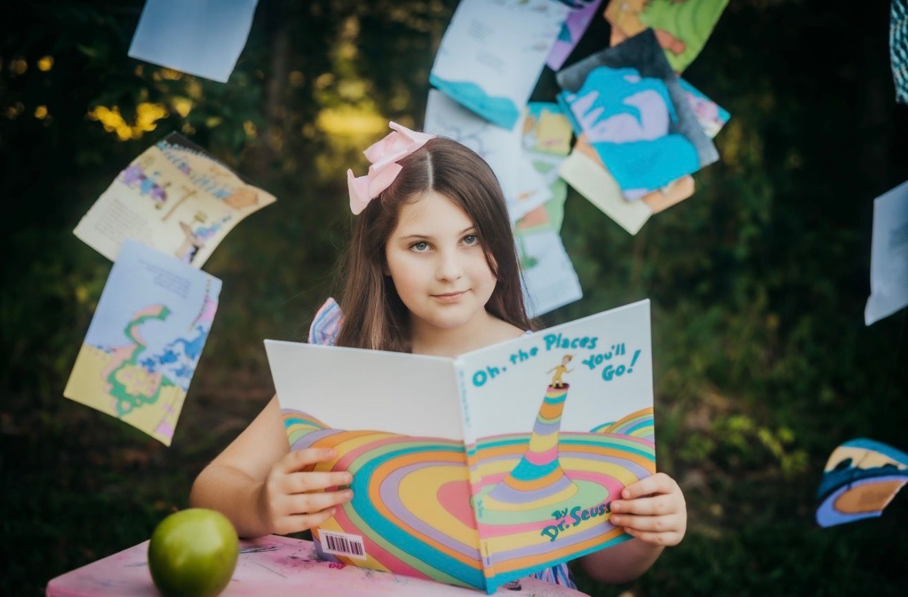 Young girl reading Dr. Seuss Oh The Places Youll Go book outdoors surrounded by flying pages.