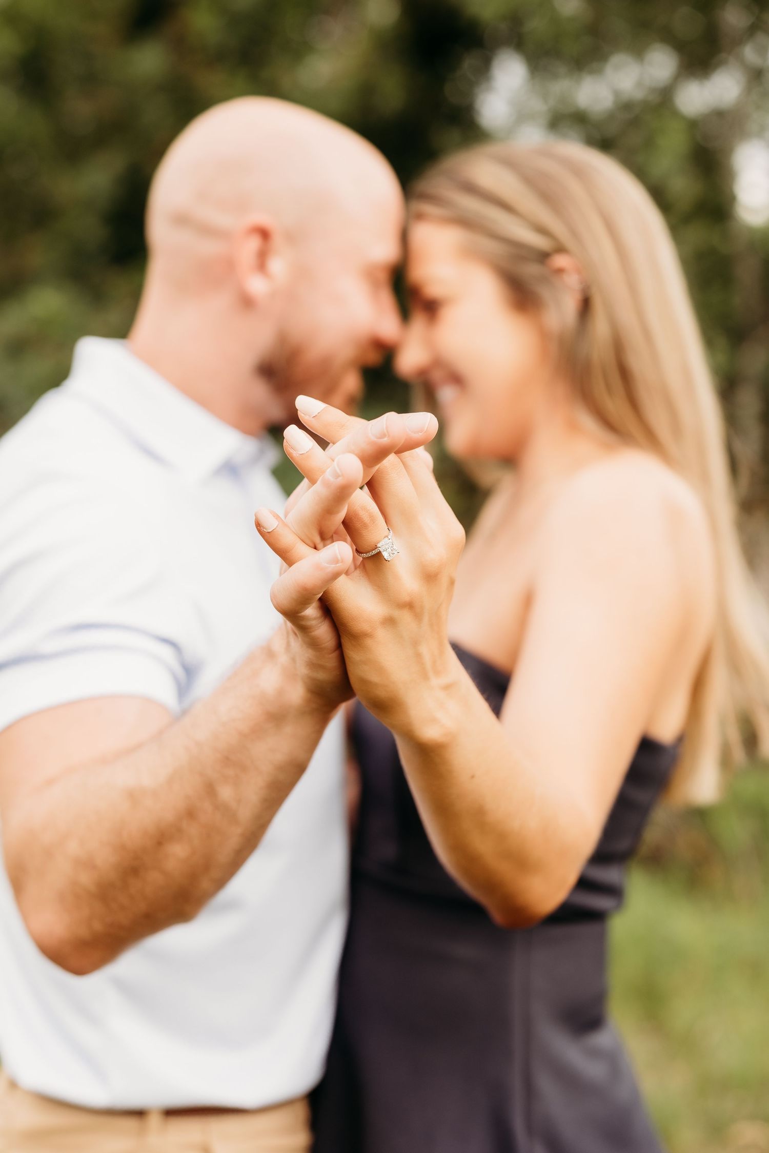 Engaged couple holds hands showing diamond ring in soft focus outdoor portrait.