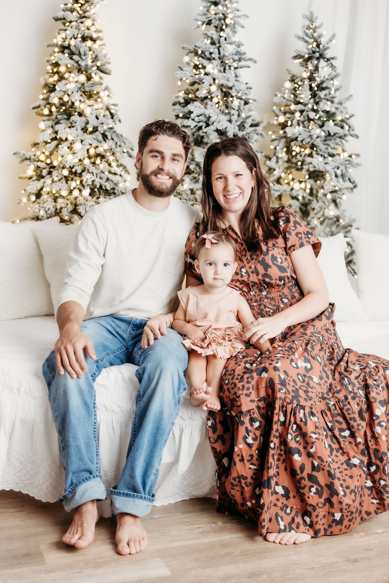 Family of three posing on white couch with snow-flocked Christmas trees, parents smiling with baby girl in festive orange dress.