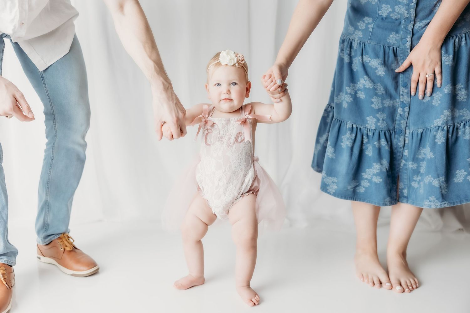 A baby girl in a pink tutu onesie takes her first steps while holding her parents hands in a bright white studio.