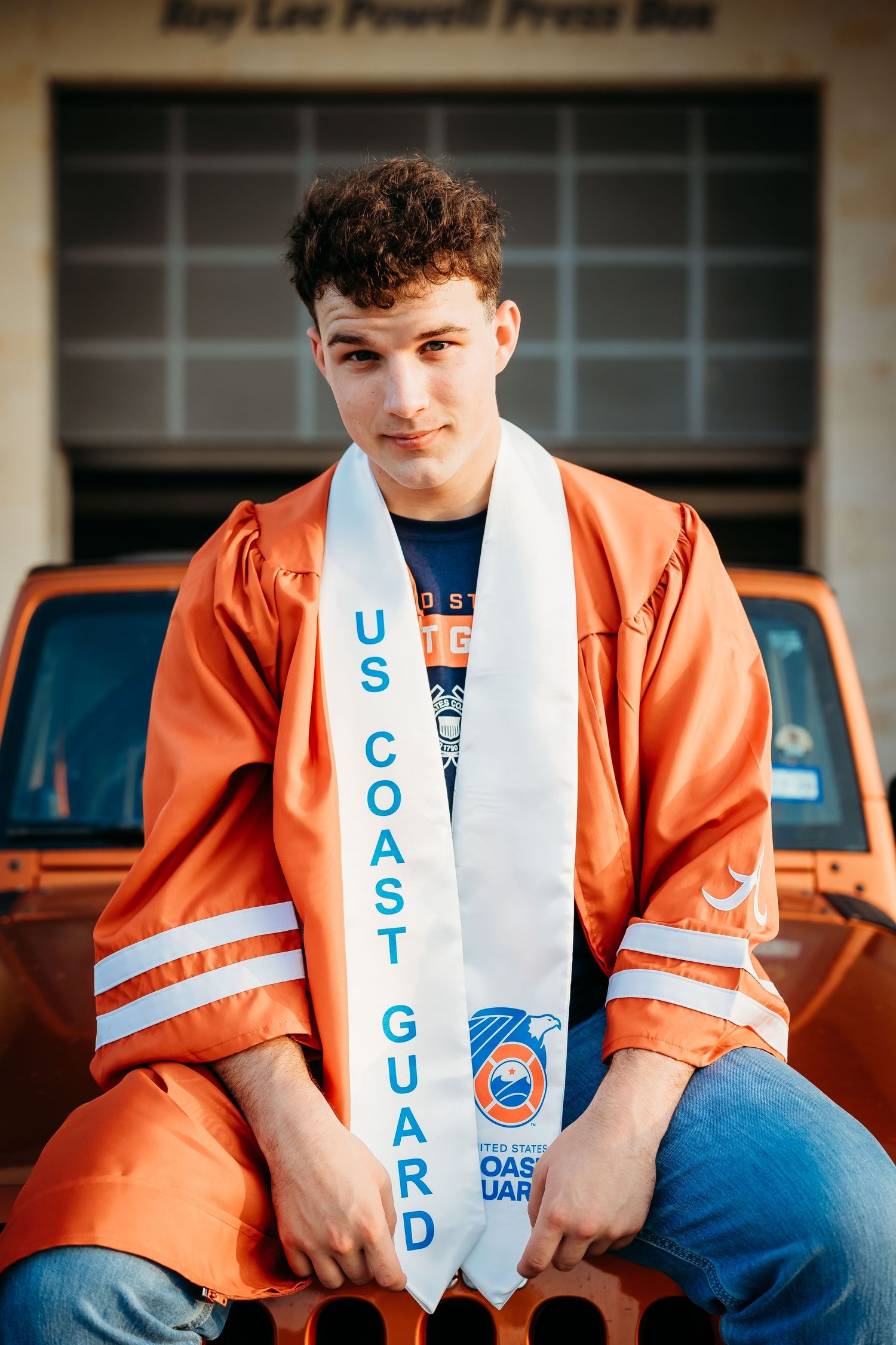 Male senior graduate in orange robe with US Coast Guard stole sits on Jeep bumper.