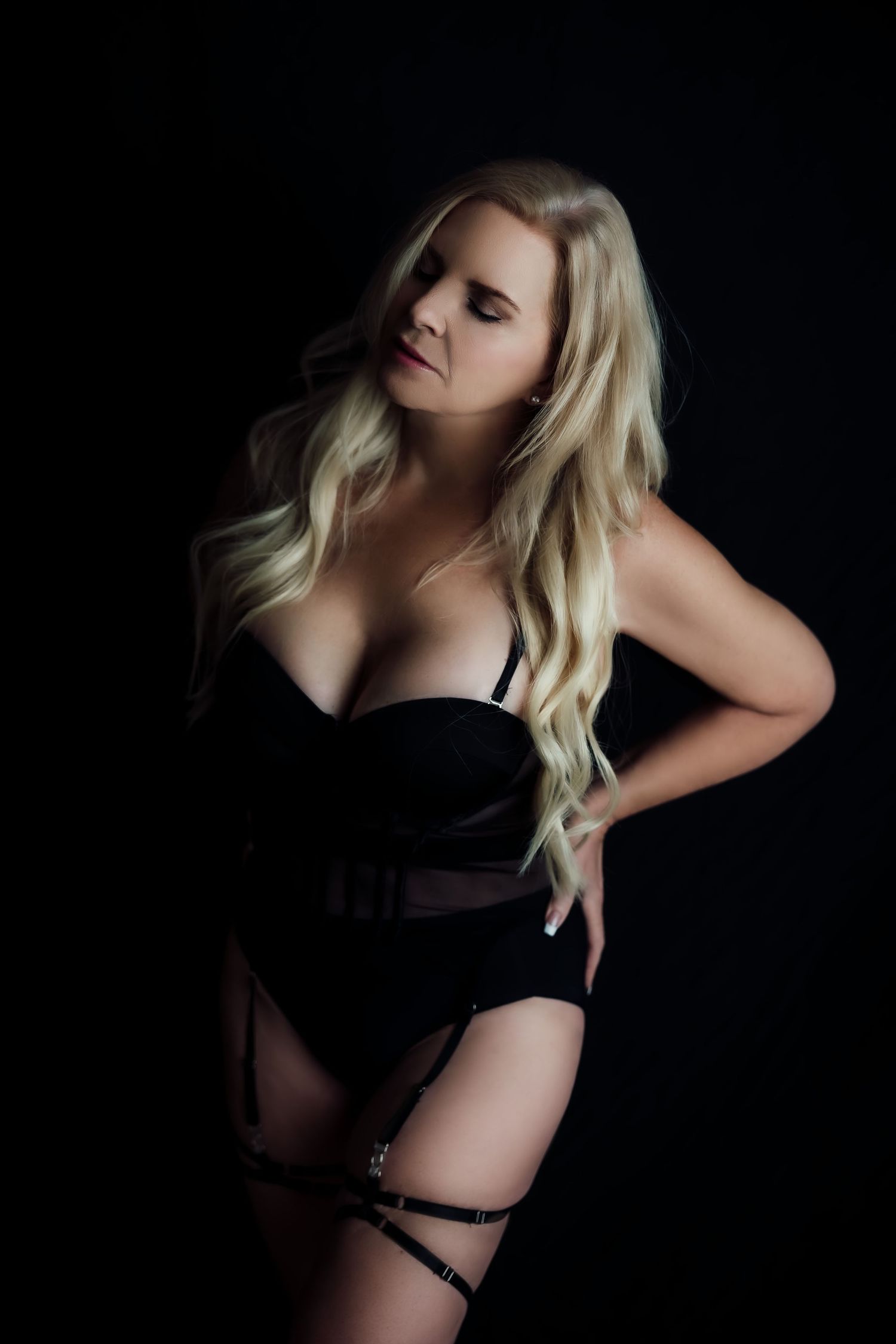 Blonde woman in black corset bodysuit and garter belt posing against dark background.