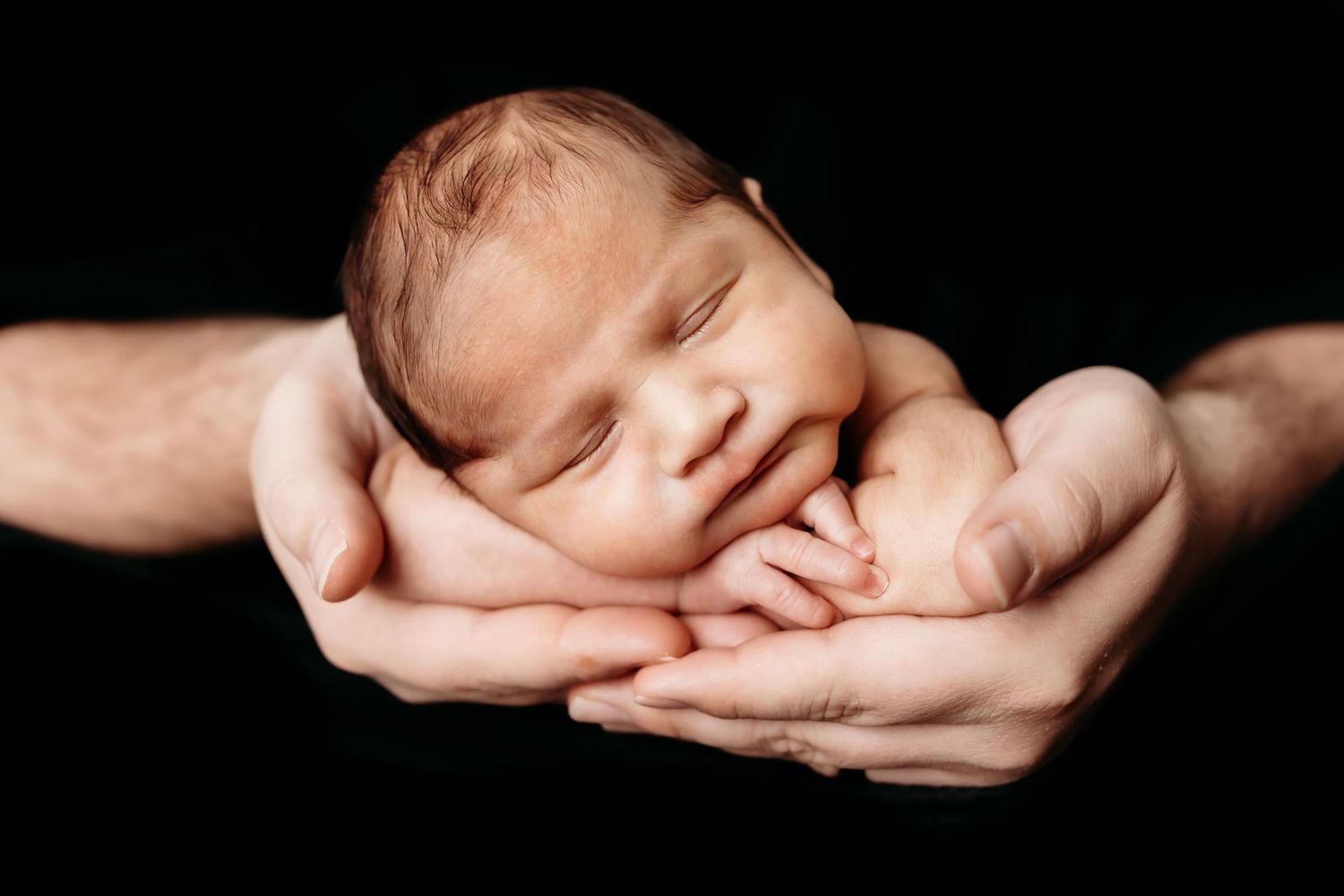 Newborn baby sleeping peacefully in parents cupped hands against a black background.