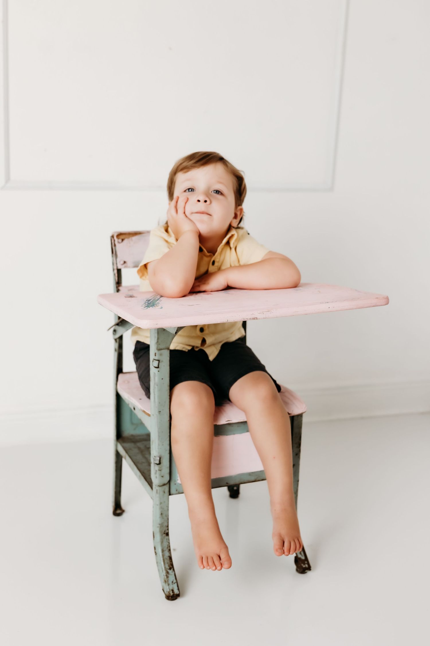 A young child sits barefoot at a vintage pink school desk, resting their cheek on their hand while gazing upward thoughtfully.