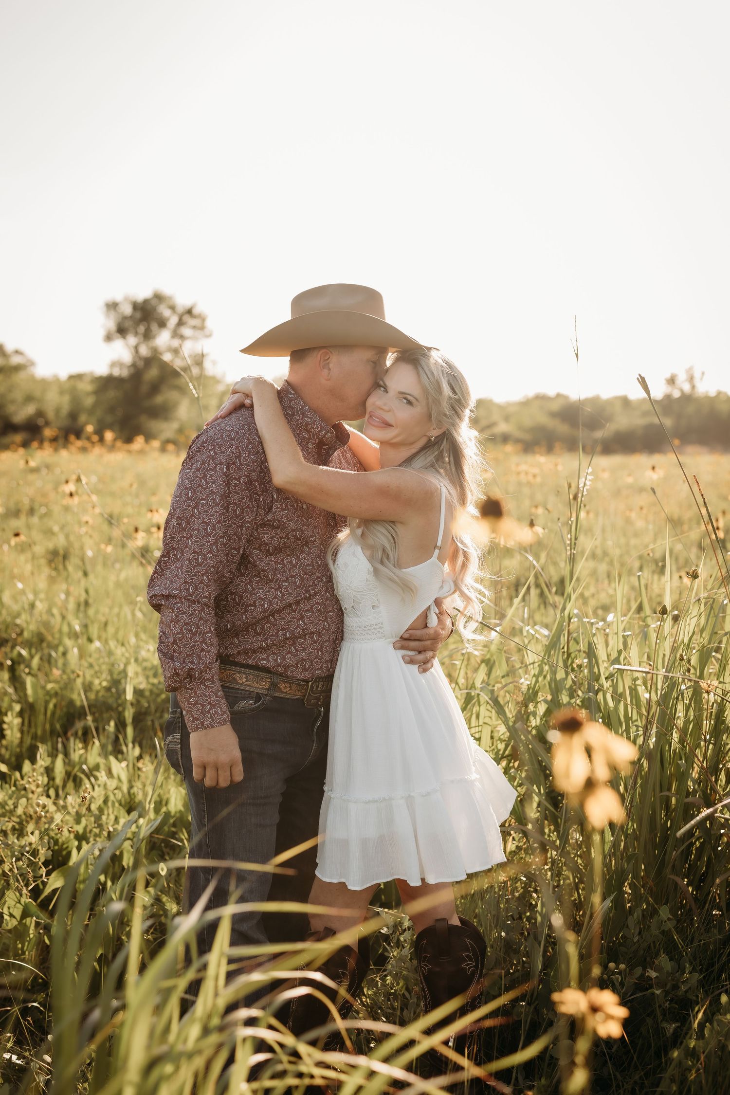 Couple embracing in golden sunflower field at sunset, man in cowboy hat kissing woman in white dress.