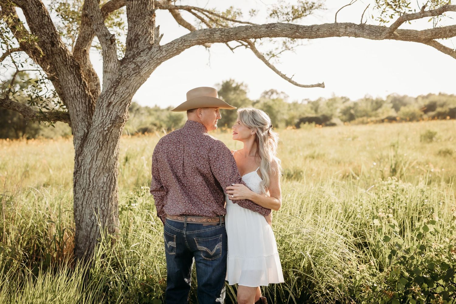 Couple embracing and gazing at each other in a lush green field beneath an oak tree.