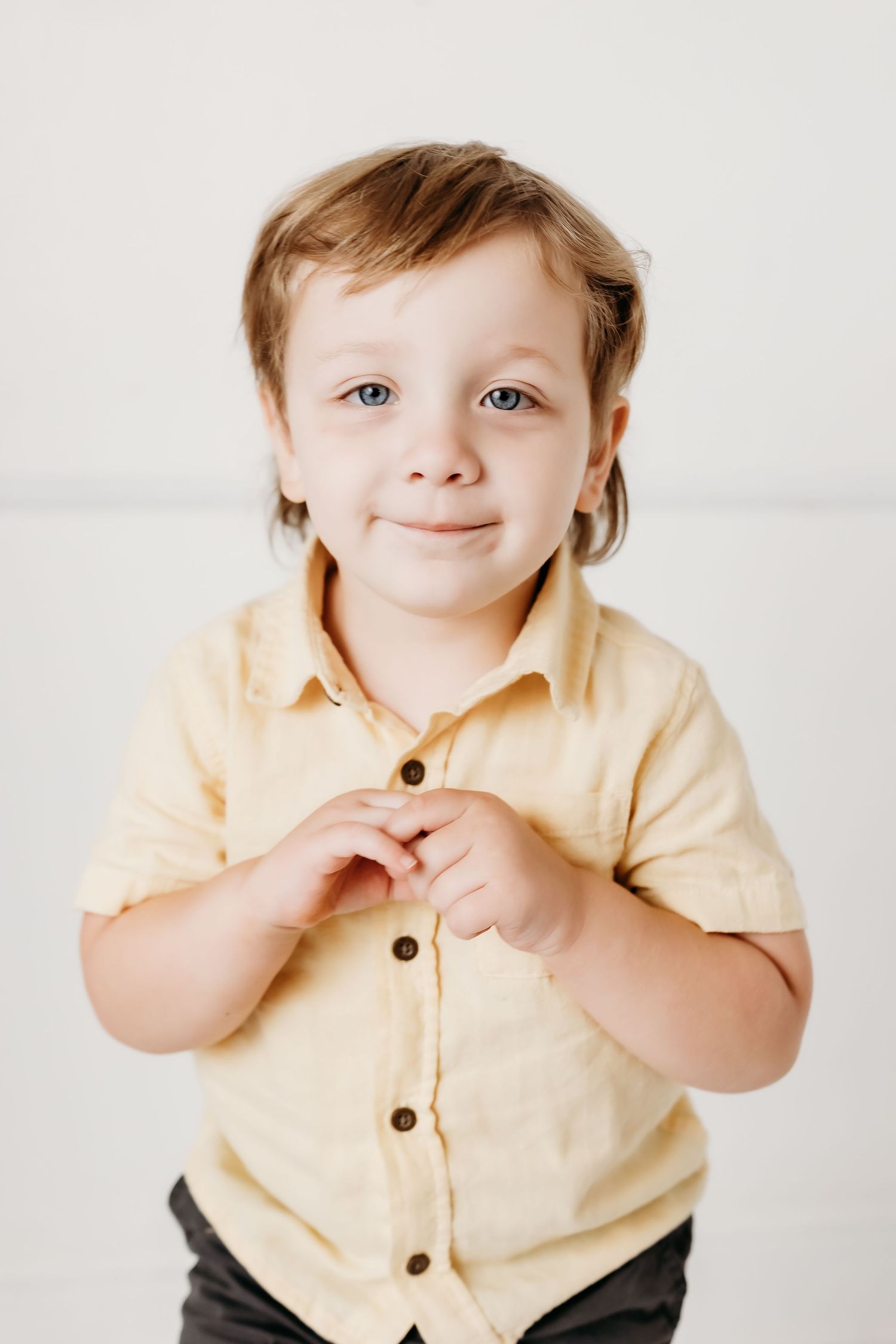 A young child with blue eyes in a yellow button-up shirt smiles warmly while standing against a white background.