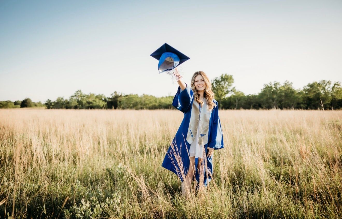 Graduate in blue cap and gown celebrates in golden field holding cap in the air.