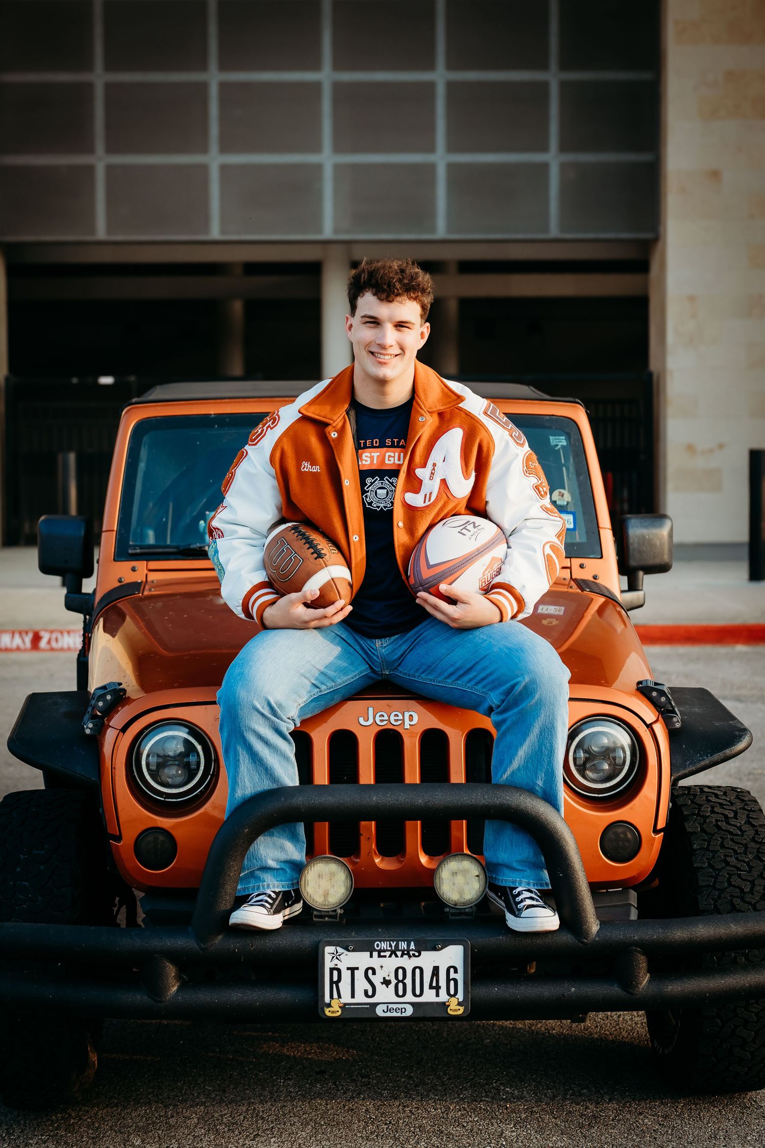 Young man in orange letterman jacket sitting on orange Jeep hood holding two footballs.