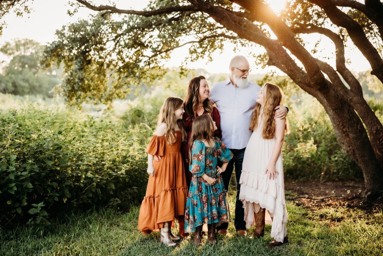 Family of five posing outdoors under a large tree in golden hour light, wearing coordinated fall outfits.