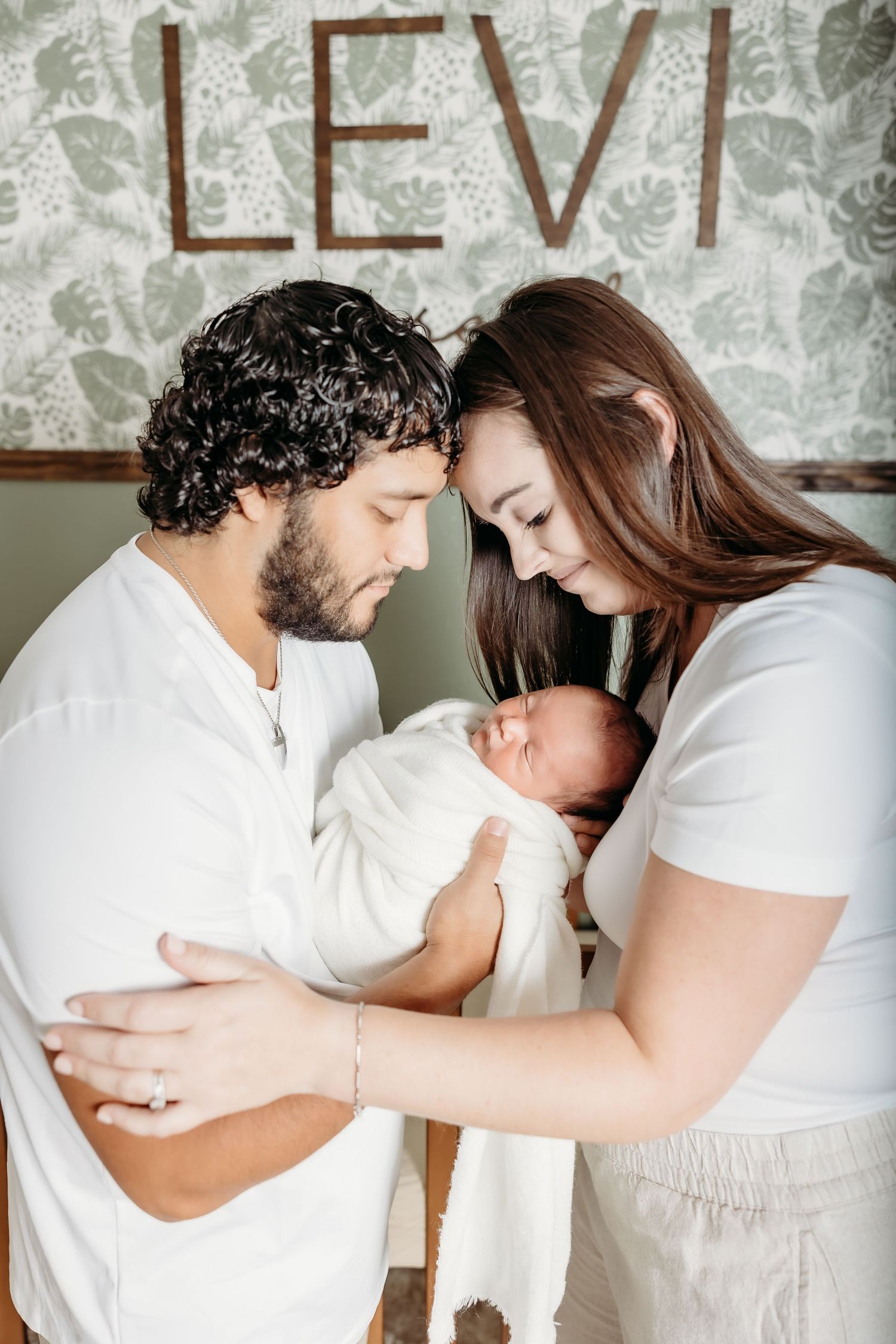 New parents in white clothing cradle swaddled newborn beneath LEVI nursery name sign.
