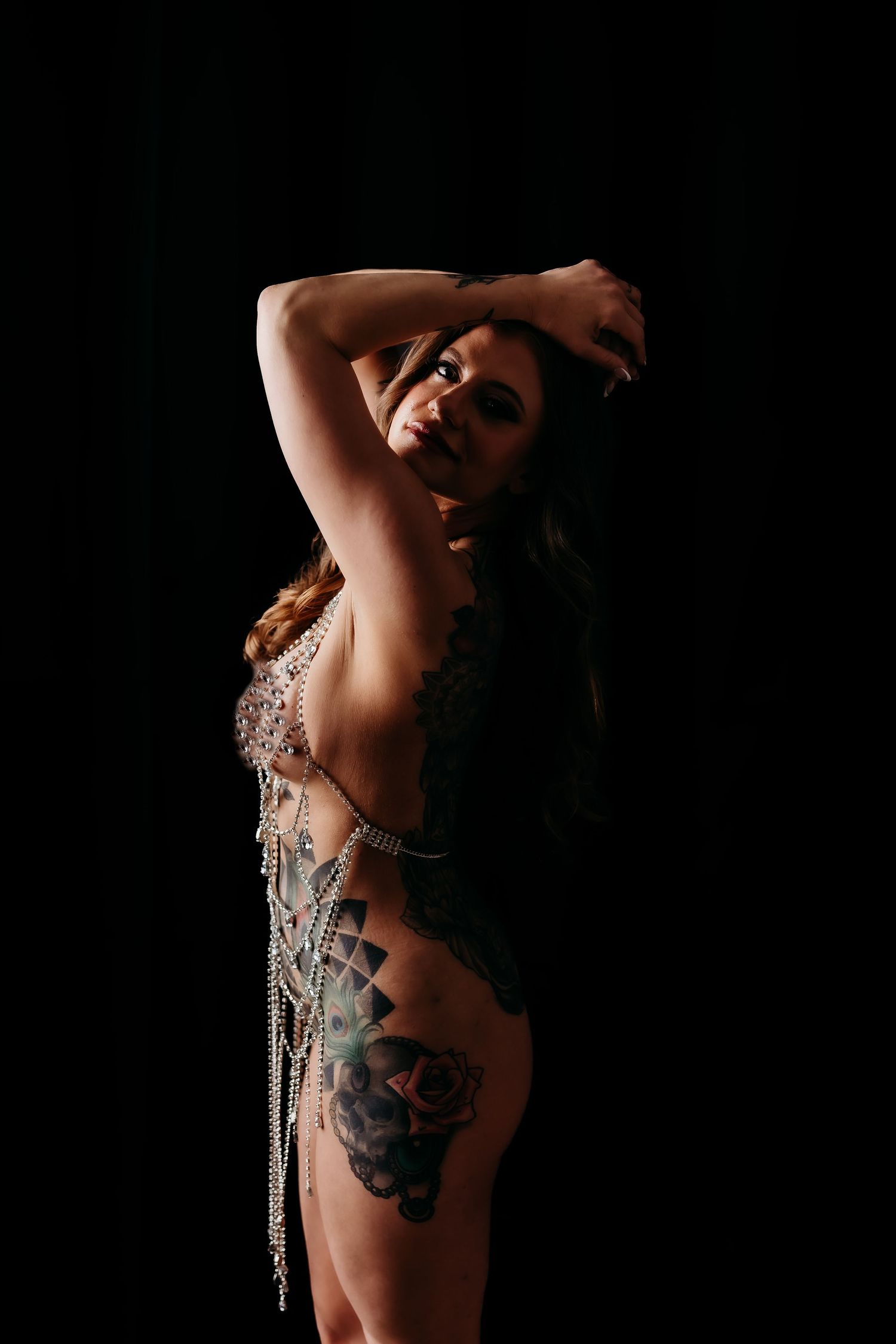 Tattooed woman posing in a jeweled bodysuit against a dark background with arms raised.
