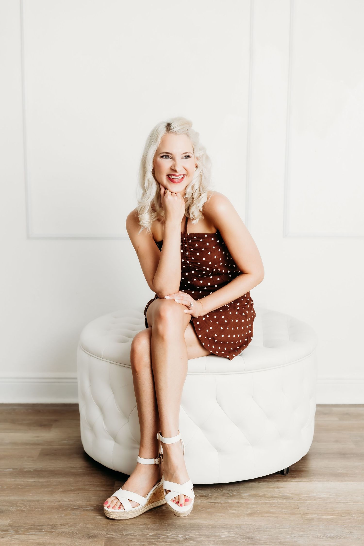 Happy blonde woman in brown polka dot dress sitting on white tufted ottoman smiling.