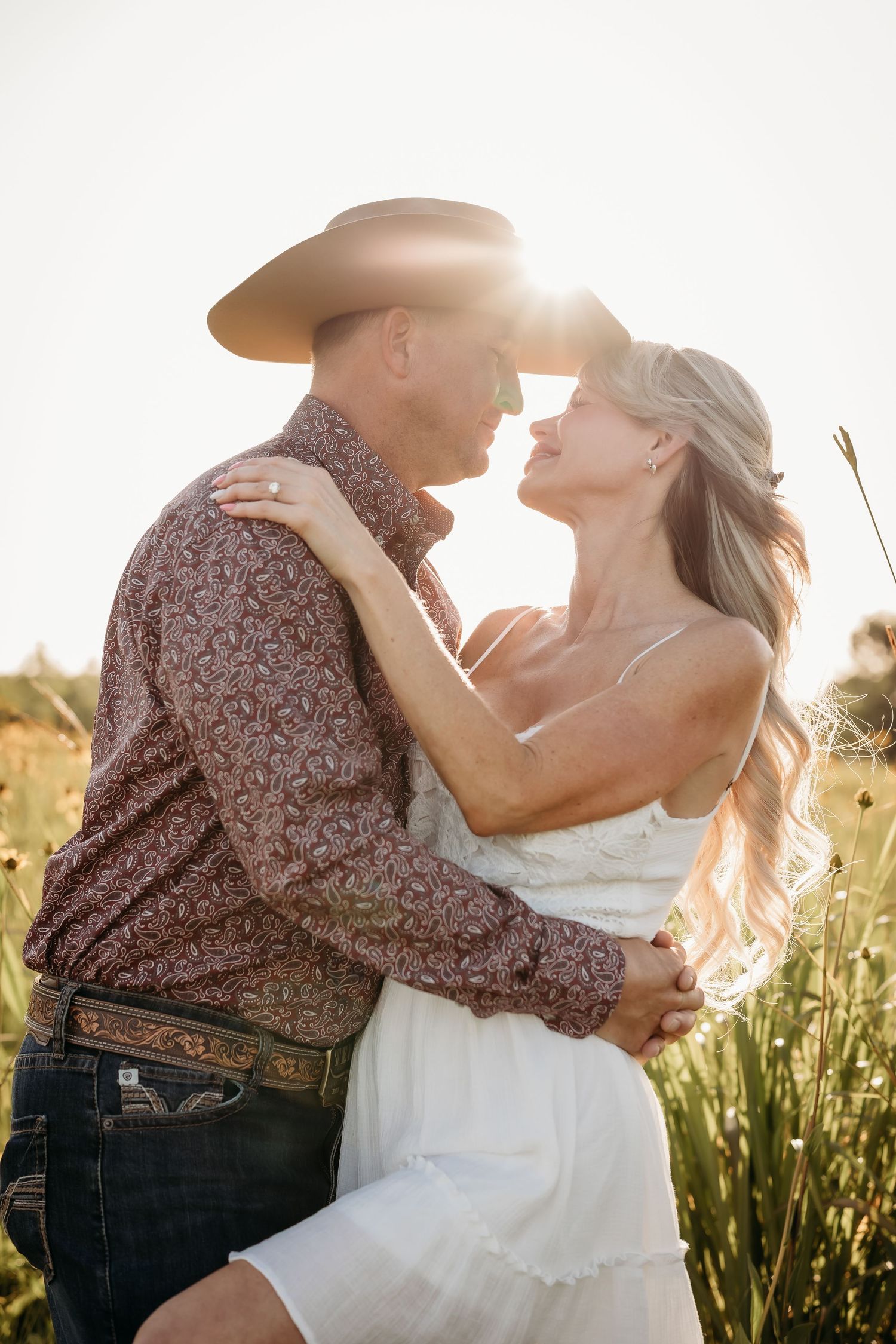 Couple in golden backlight embracing closely in a wildflower field, man wearing a cowboy hat.
