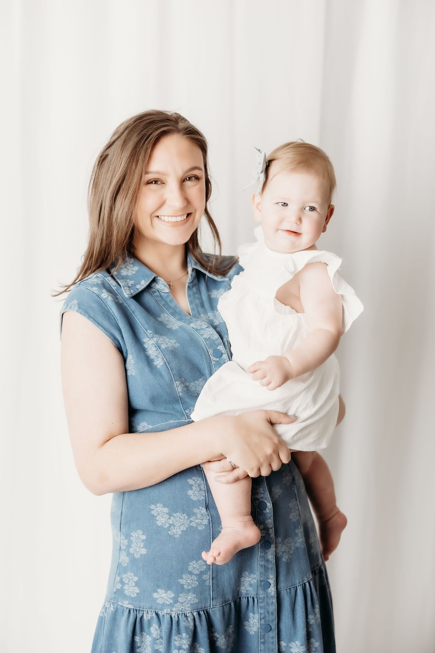 Smiling woman in blue floral dress holding a happy baby girl in white dress against white curtain backdrop.