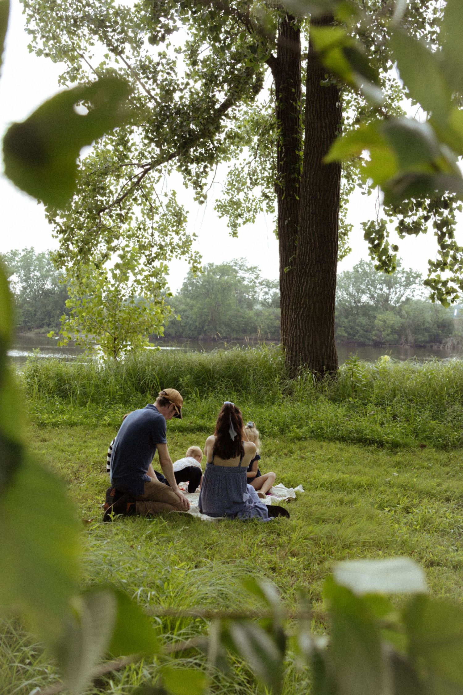 Family relaxing together on grass near a tree by a river during an outdoor picnic.