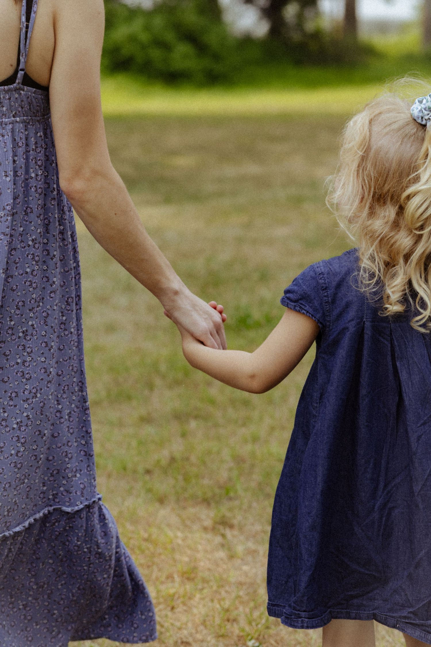 A mother in a purple floral dress holds hands with her young blonde daughter in a blue dress outdoors.
