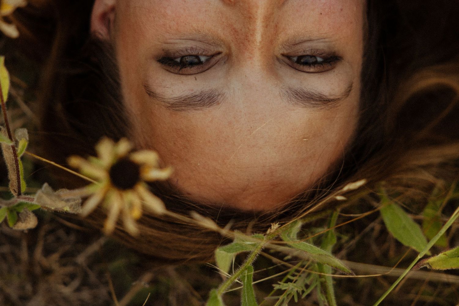 Close-up of a woman lying in grass with closed eyes and freckles, surrounded by wildflowers.