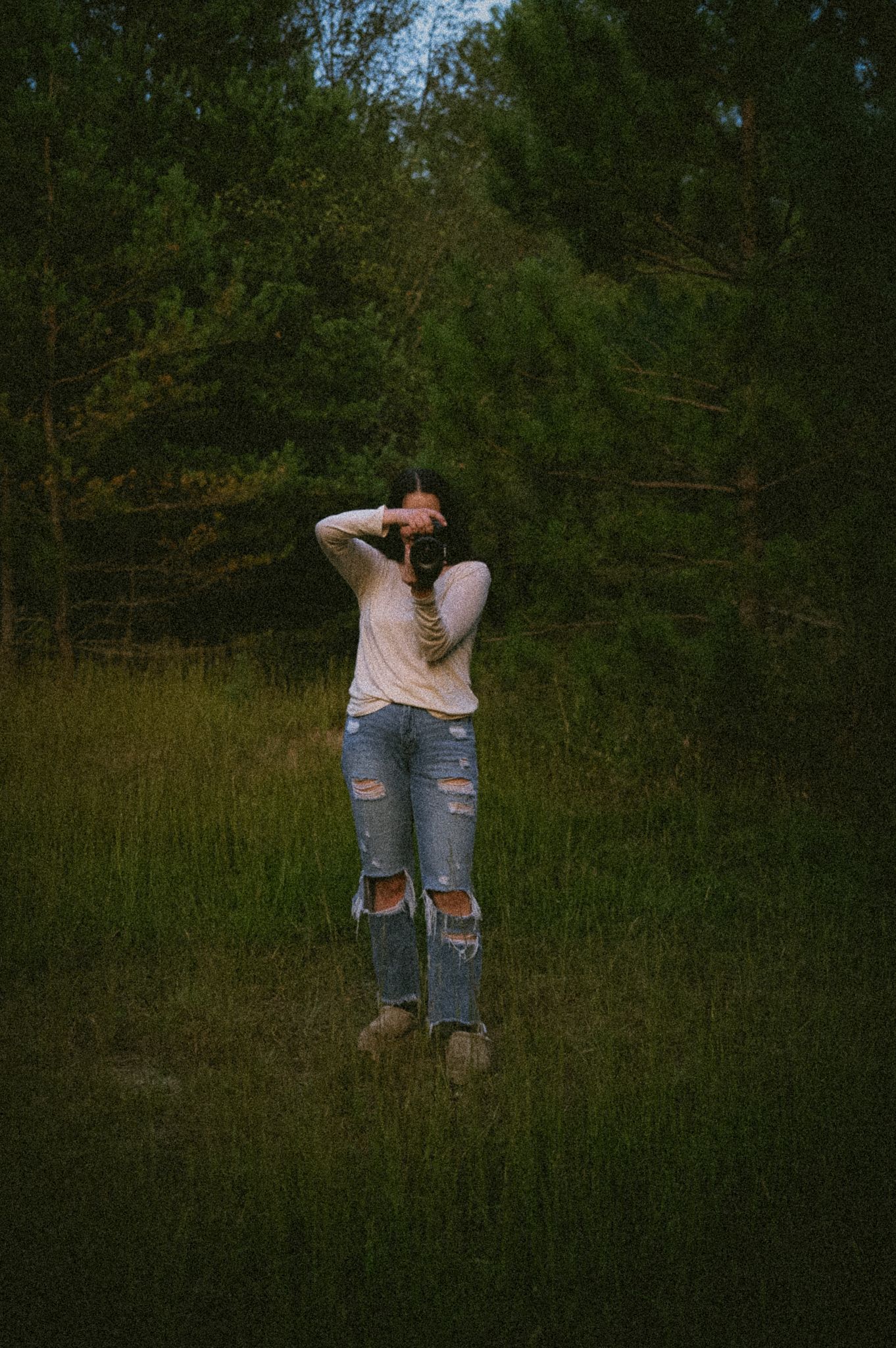 Woman in ripped jeans and white top holds camera to face while standing in a lush green forest meadow.