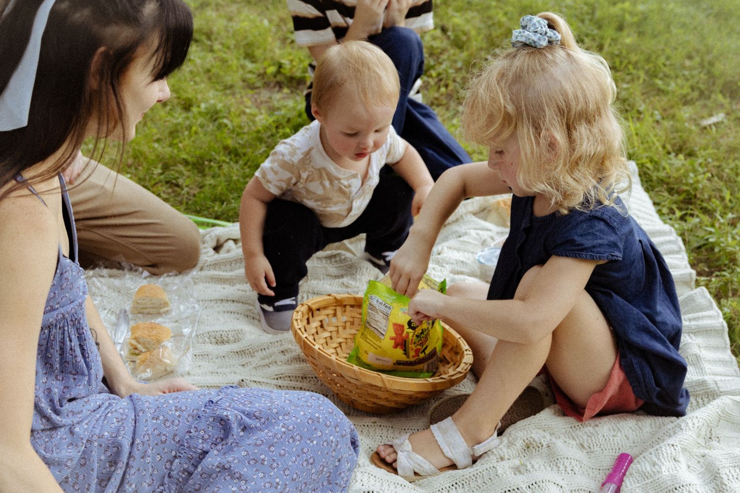 Children and a woman enjoy a sunny outdoor picnic on a blanket with a wicker basket full of snacks.
