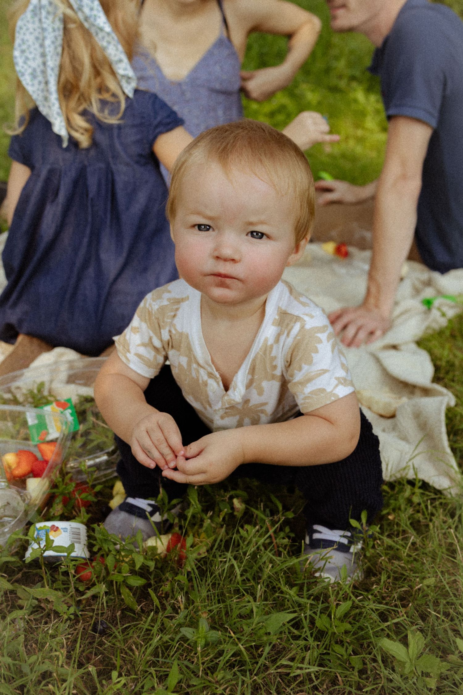 A curious toddler sits on grass during a family picnic, wearing a patterned shirt, with adults relaxing on a blanket behind.