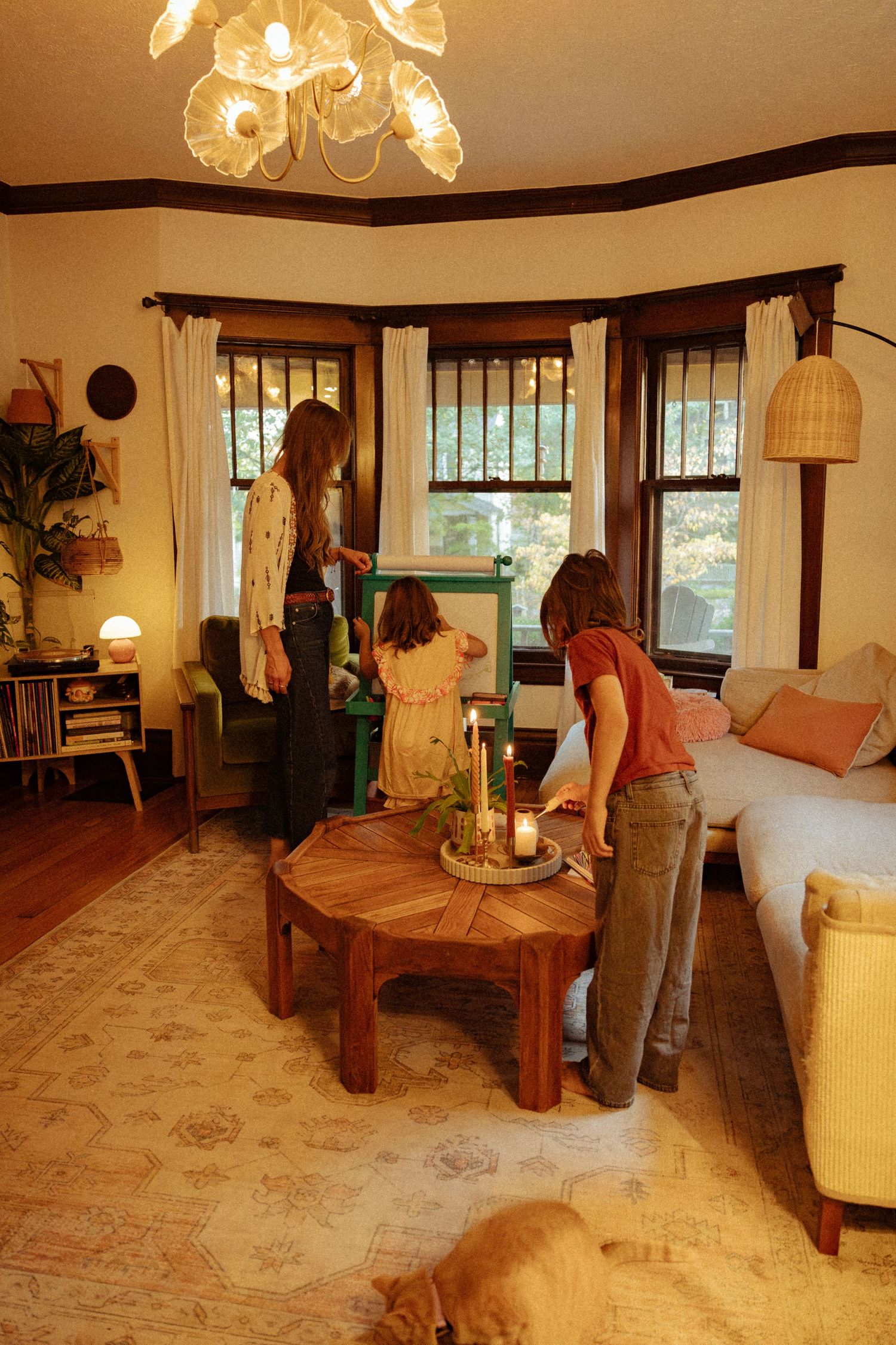 A woman and two children gather around a wooden coffee table with candles in a cozy living room with a bay window and chandelier.