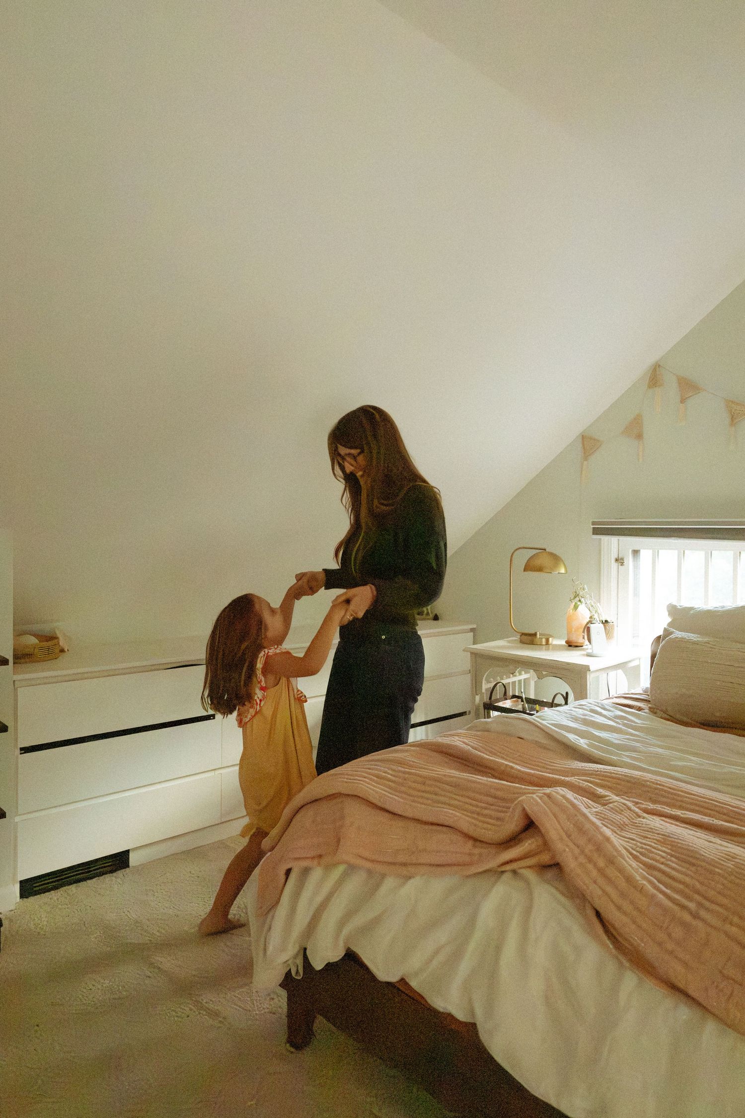 Mother and young daughter in a yellow dress hold hands and dance in a bright attic bedroom with a cozy bed and pennant garland.