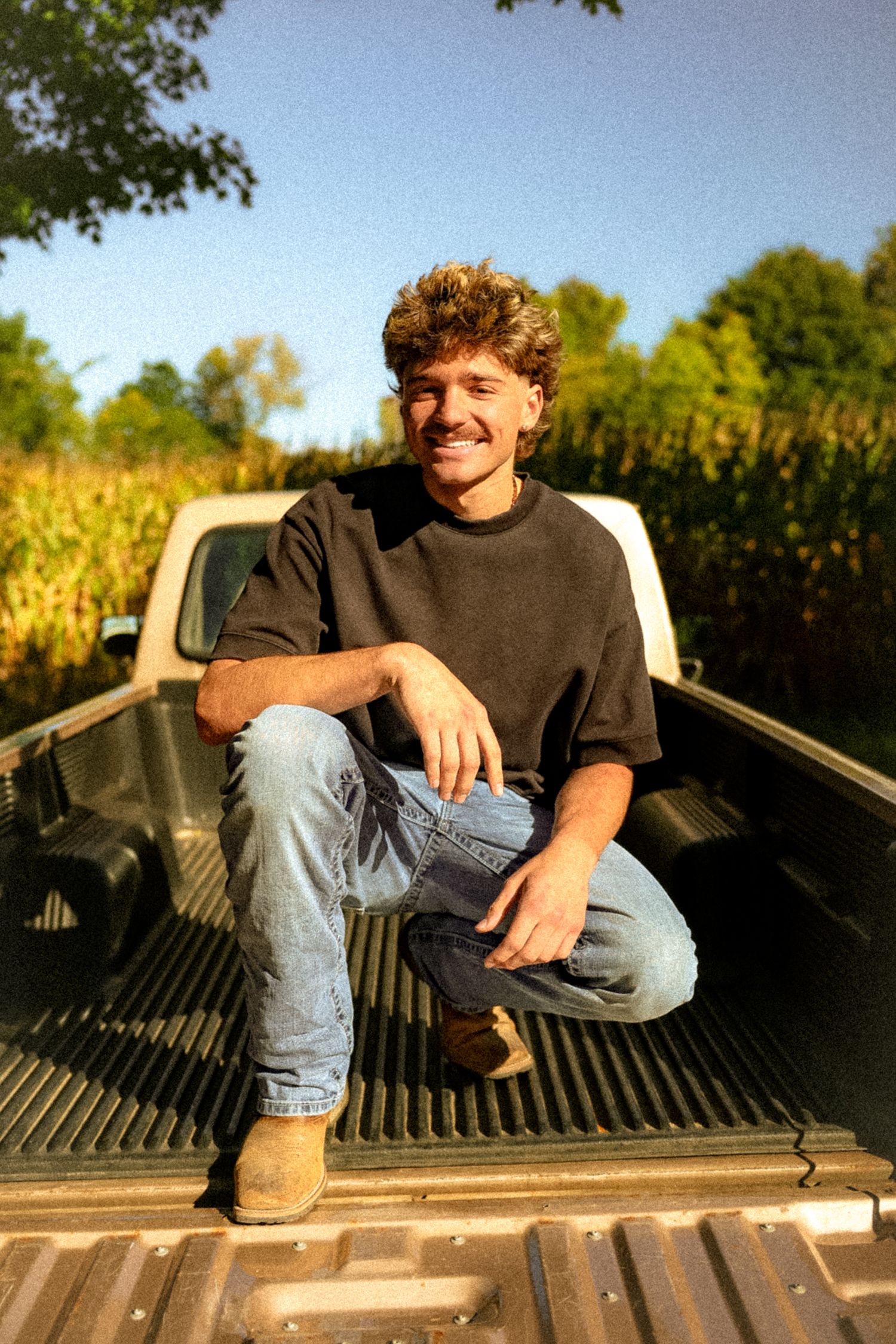 Smiling young man with curly hair sitting in a truck bed near a cornfield, wearing jeans and a black t-shirt.