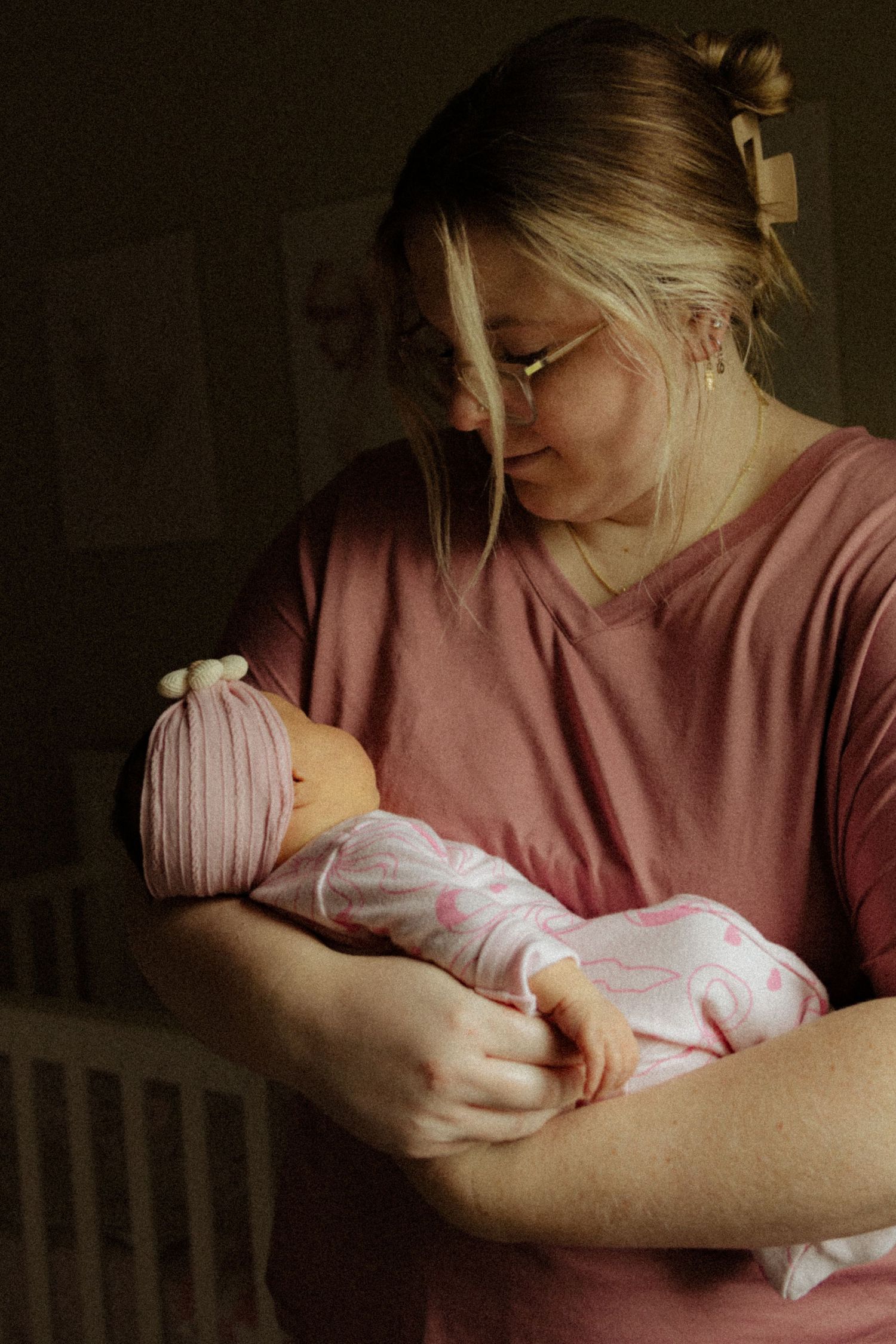 Mother in pink scrubs tenderly holding her newborn baby dressed in pink onesie and striped hat.