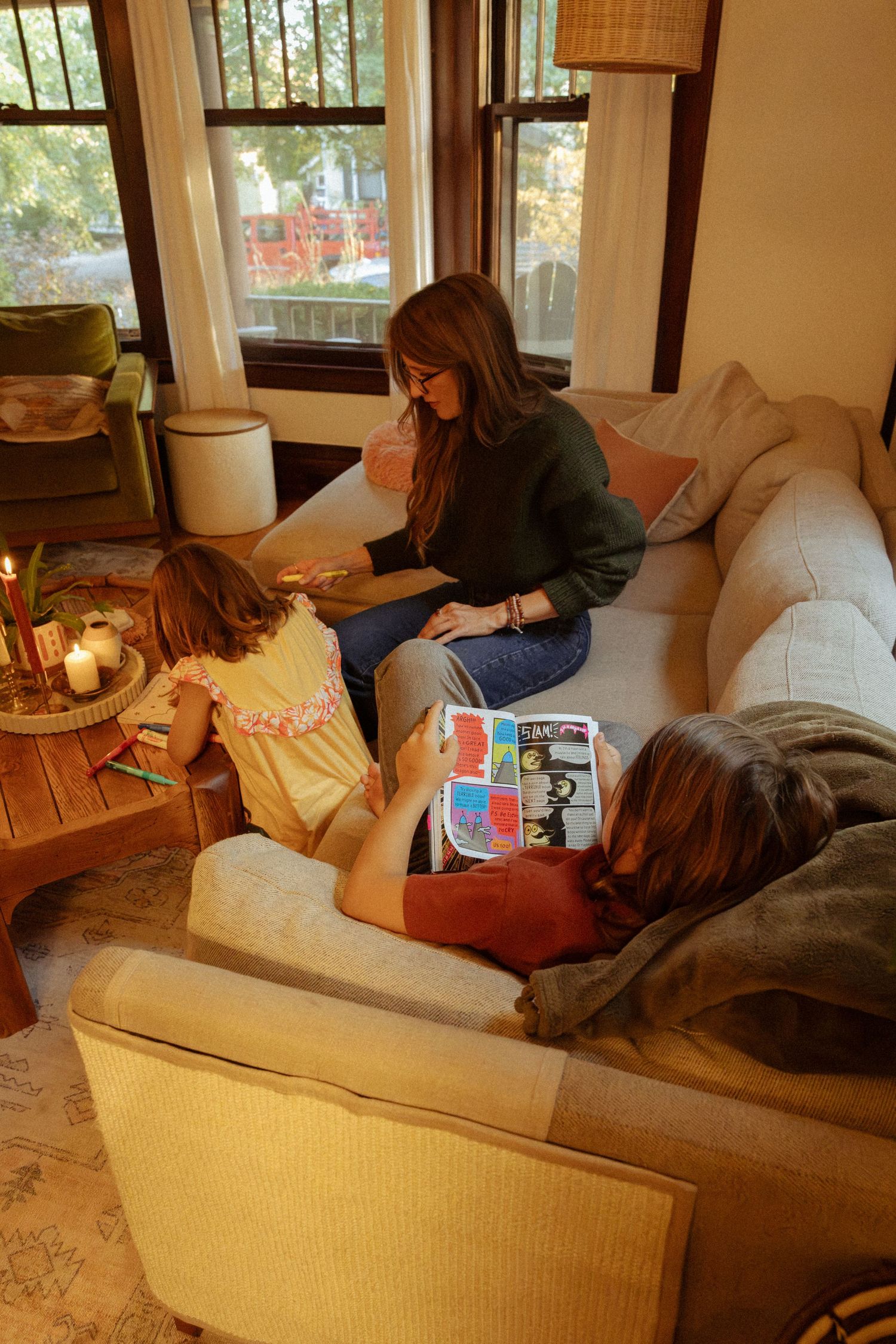 A woman relaxes on a cream sofa while a child colors at a wooden table and another reads a colorful book nearby.